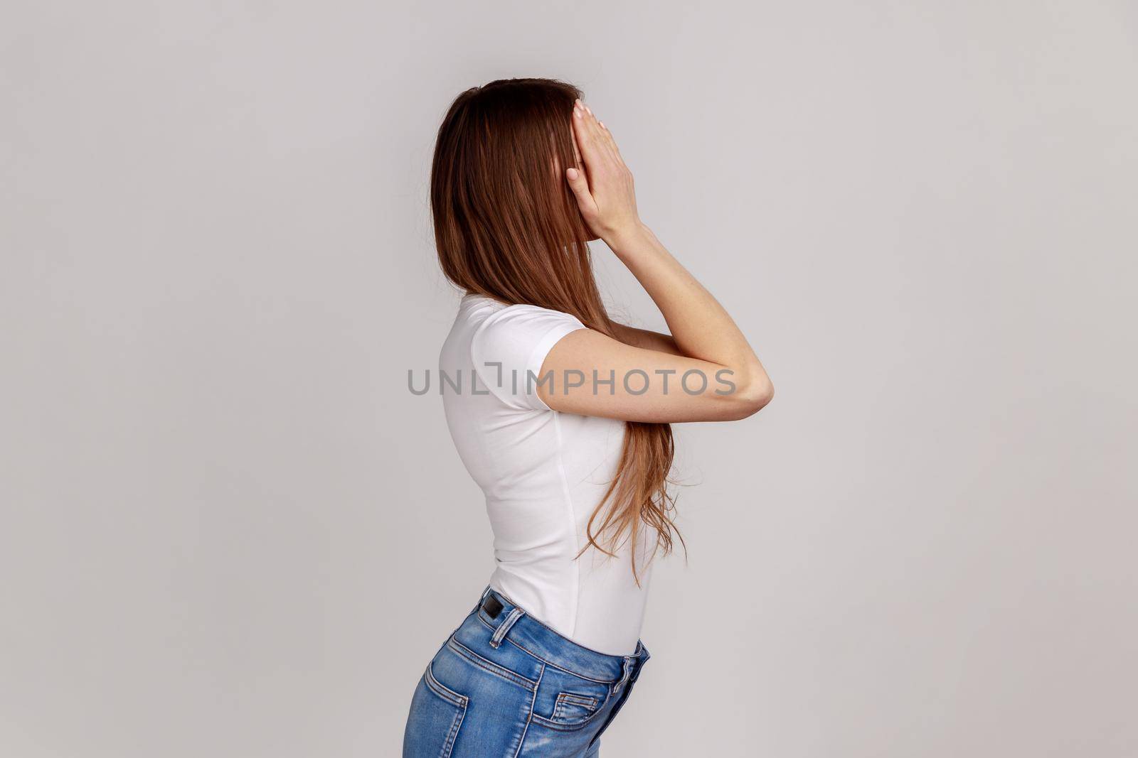 Side view portrait of young adult dark haired woman standing, covering eyes with hand, refusing to watch, shame content, wearing white T-shirt. Indoor studio shot isolated on gray background.