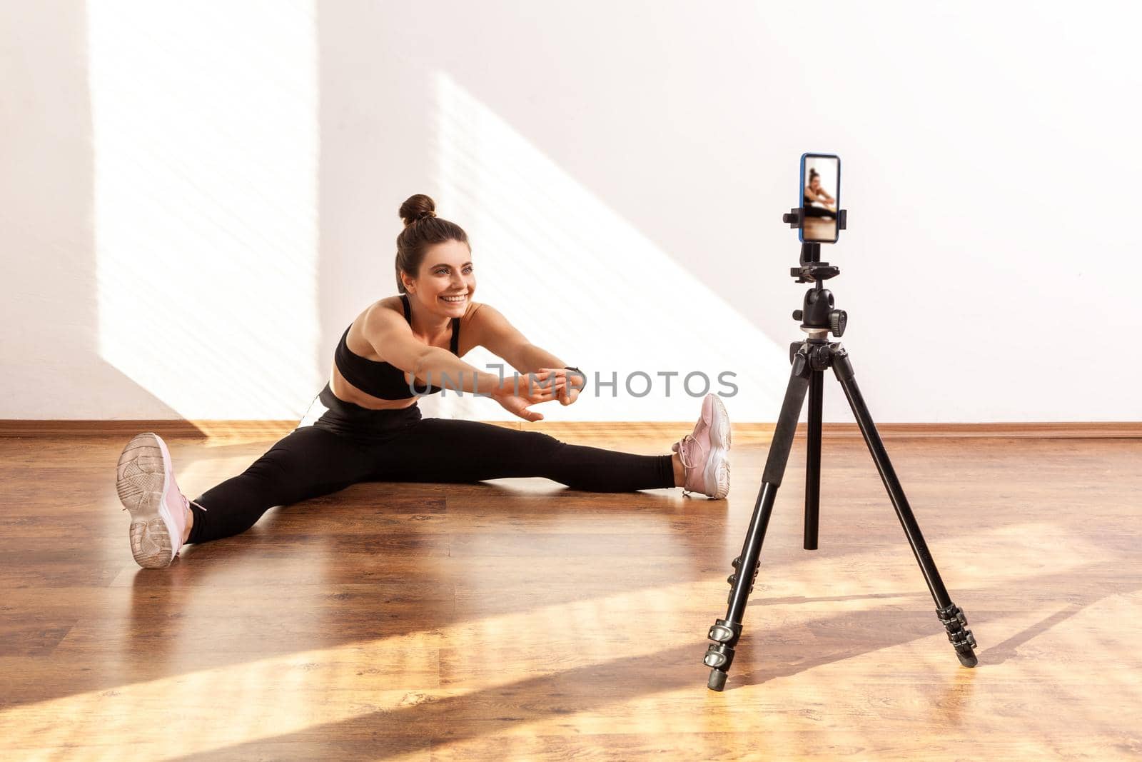Female online trainer using phone on tripod for recording tutorials of stretching after workout, wearing black sports top and tights. Full length studio shot illuminated by sunlight from window.