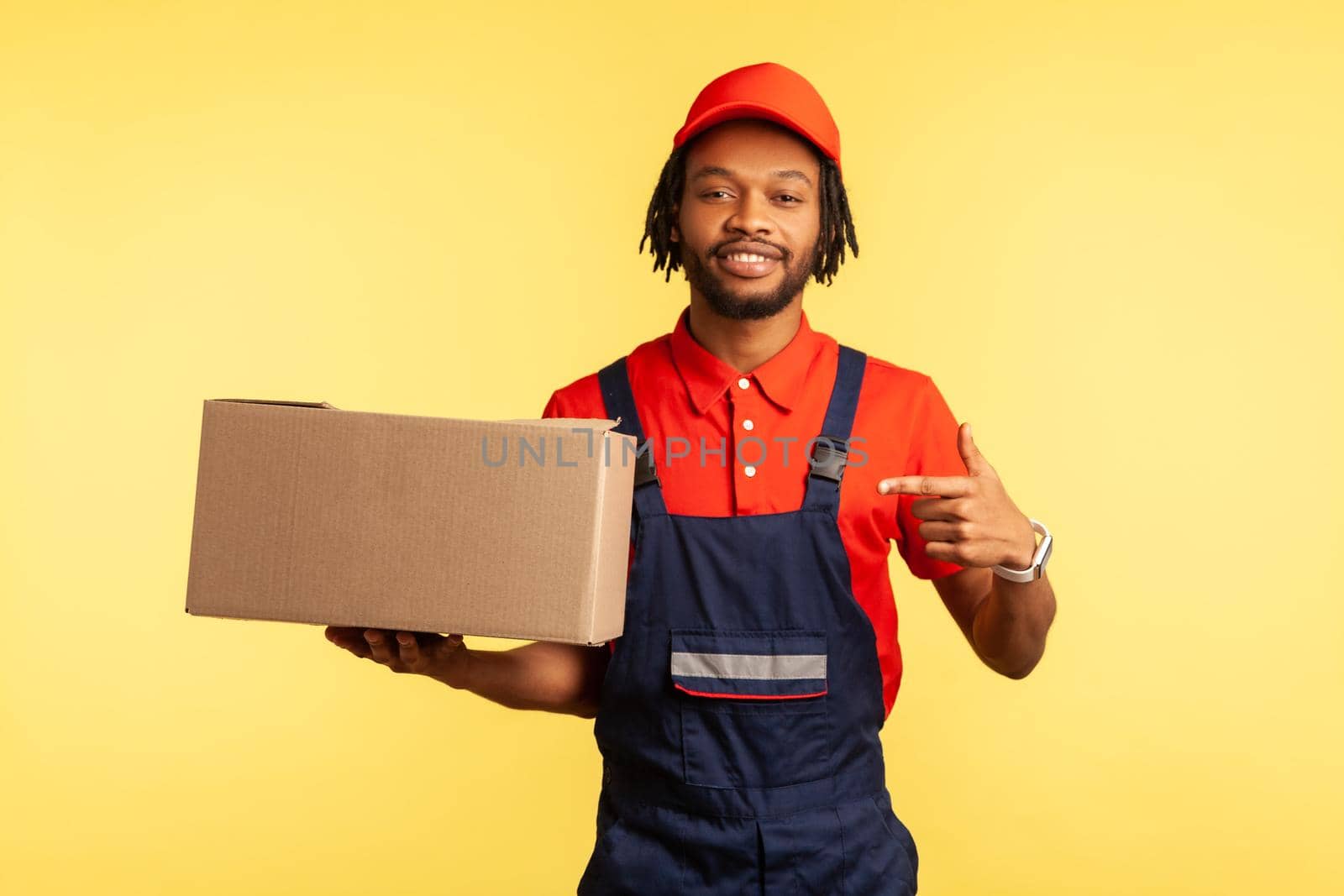 Portrait of cargo transportation, delivery service. Friendly courier in uniform pointing at cardboard box, delivering parcel to client. Indoor studio shot isolated on yellow background.