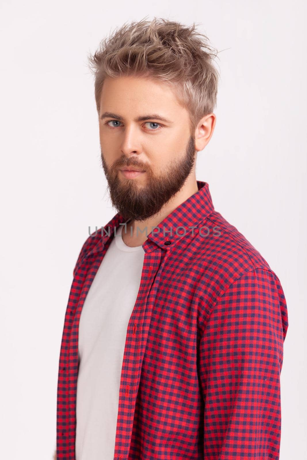 Portrait of young self-assured bearded man in plaid t-shirt confidently looking at camera, pick-up tricks. Indoor studio shot isolated on gray background
