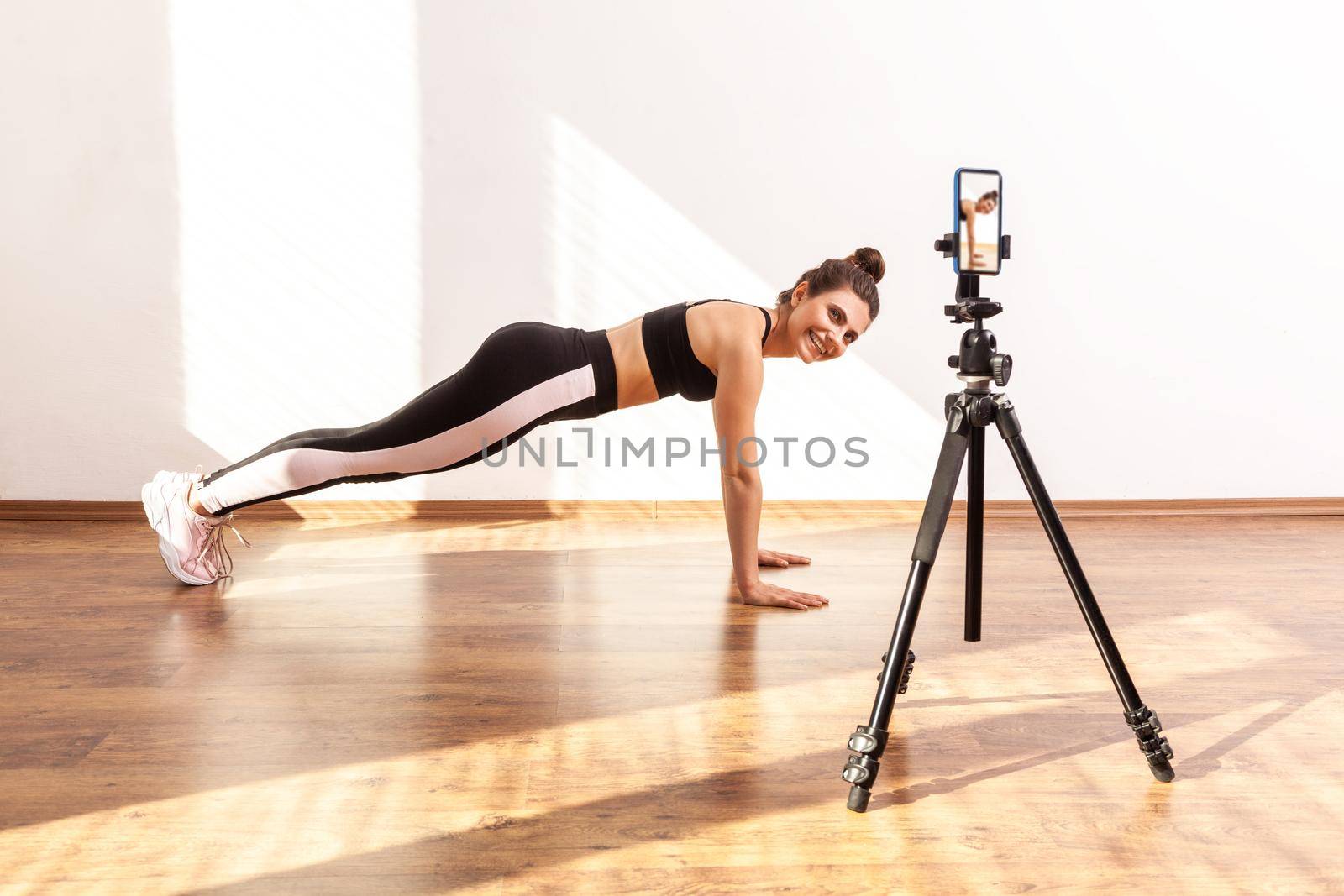 Coach teaching correct technique of doing plank exercise online, recording video on phone camera, wearing black sports top and tights. Full length studio shot illuminated by sunlight from window.