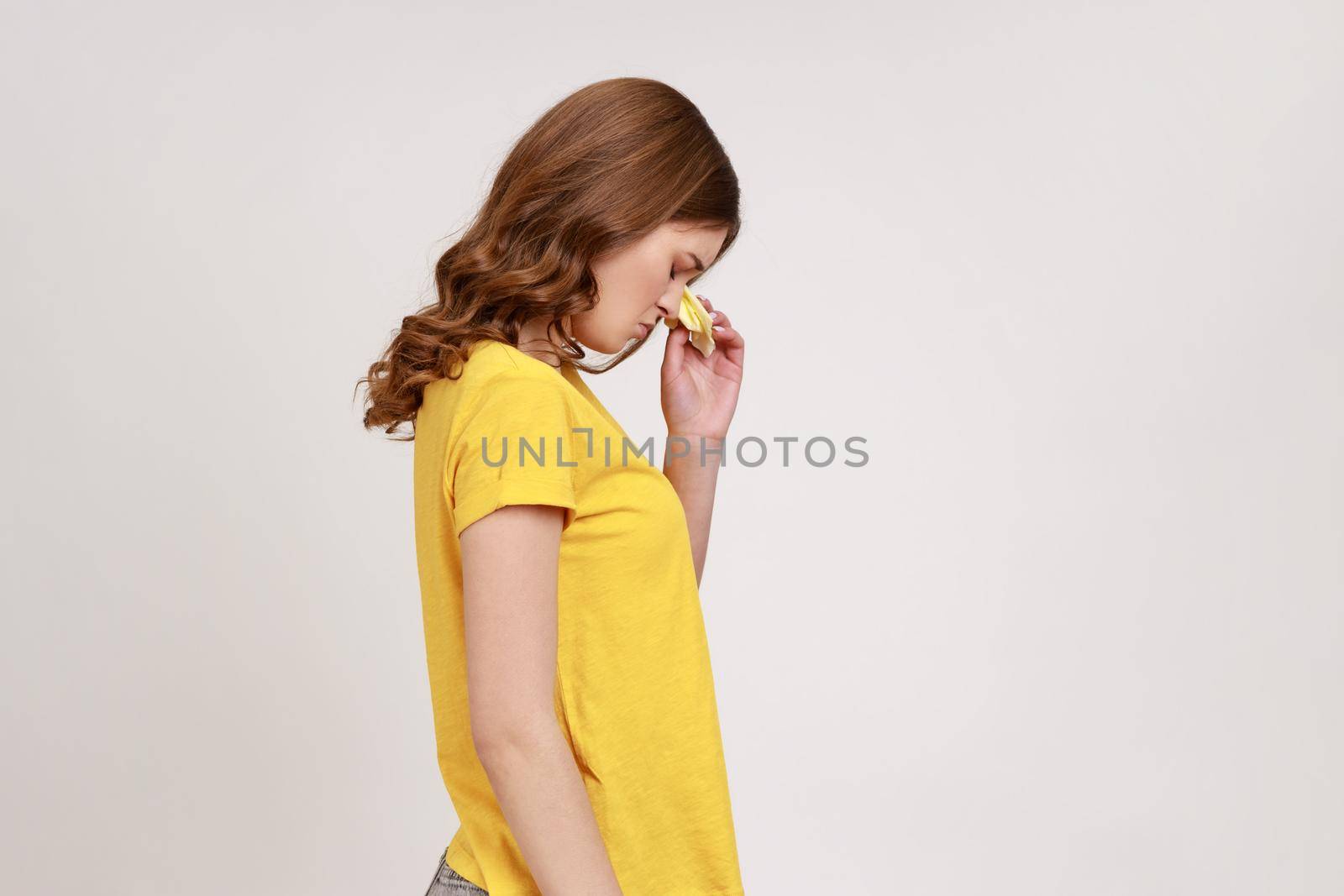 Profile portrait of teenager girl with wavy hair in yellow T-shirt expressing sadness and depression, upset female crying, desperate sorrowful feelings. Indoor studio shot isolated on gray background.