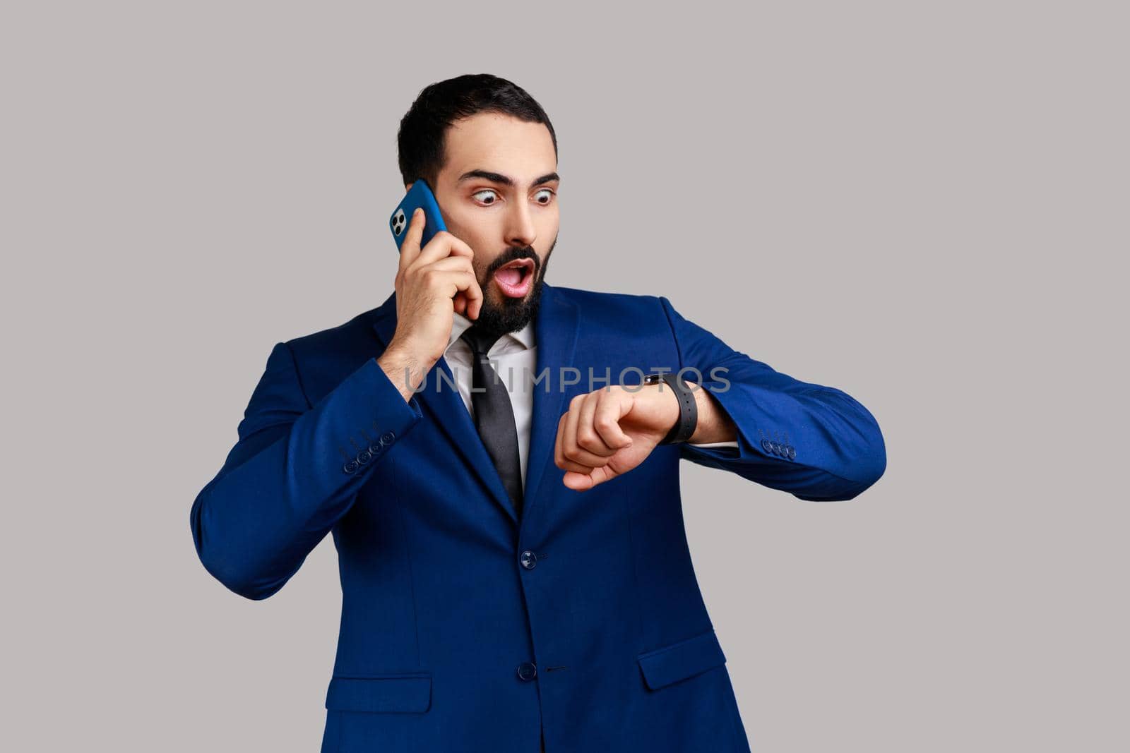 Portrait of handsome bearded man talking phone and looking at wristwatch, being late for date or business meeting, wearing official style suit. Indoor studio shot isolated on gray background.