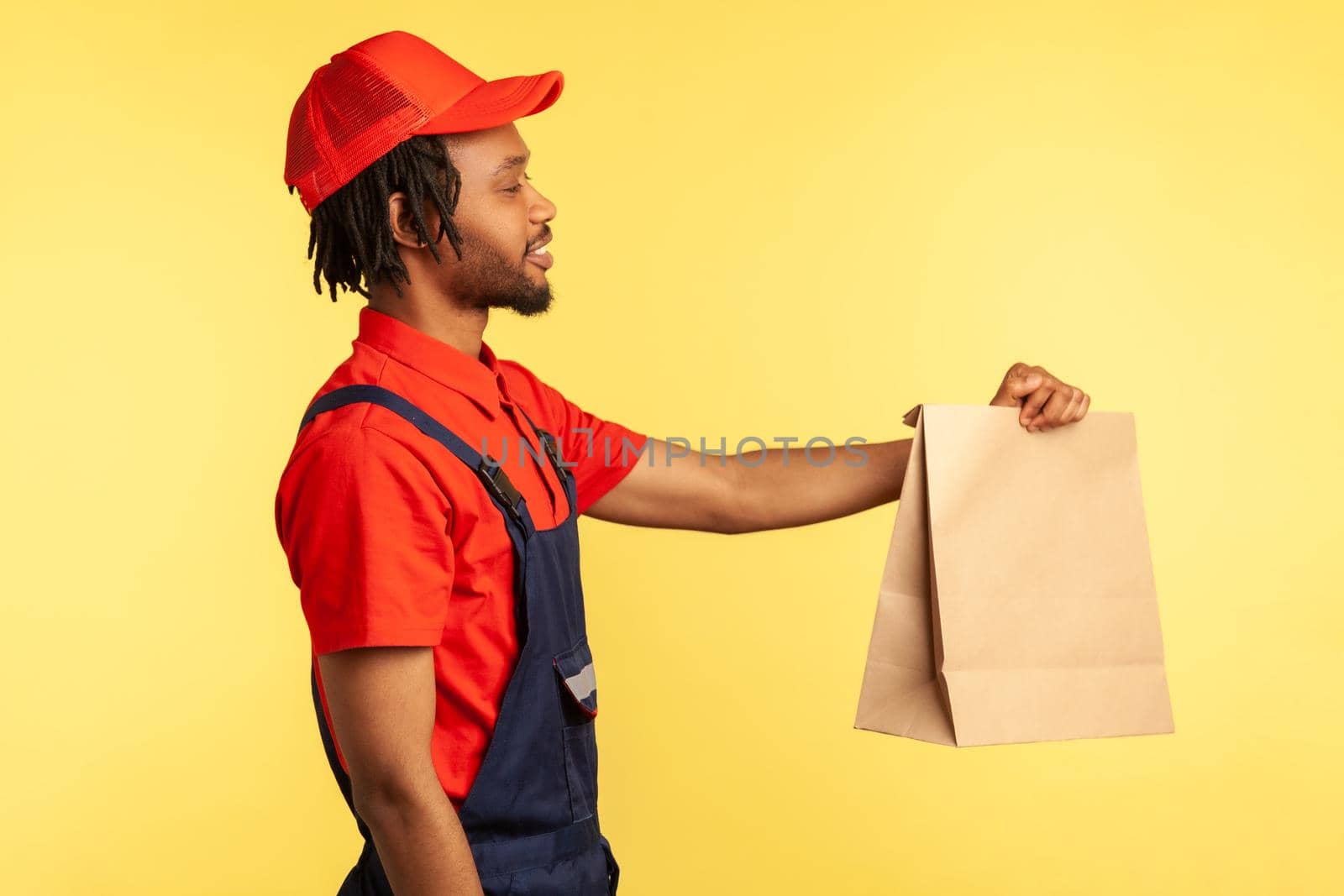 Side view portrait of bearded deliveryman wearing red visor cap and blue overalls, giving paper parcel to client, fast delivery service. Indoor studio shot isolated on yellow background.