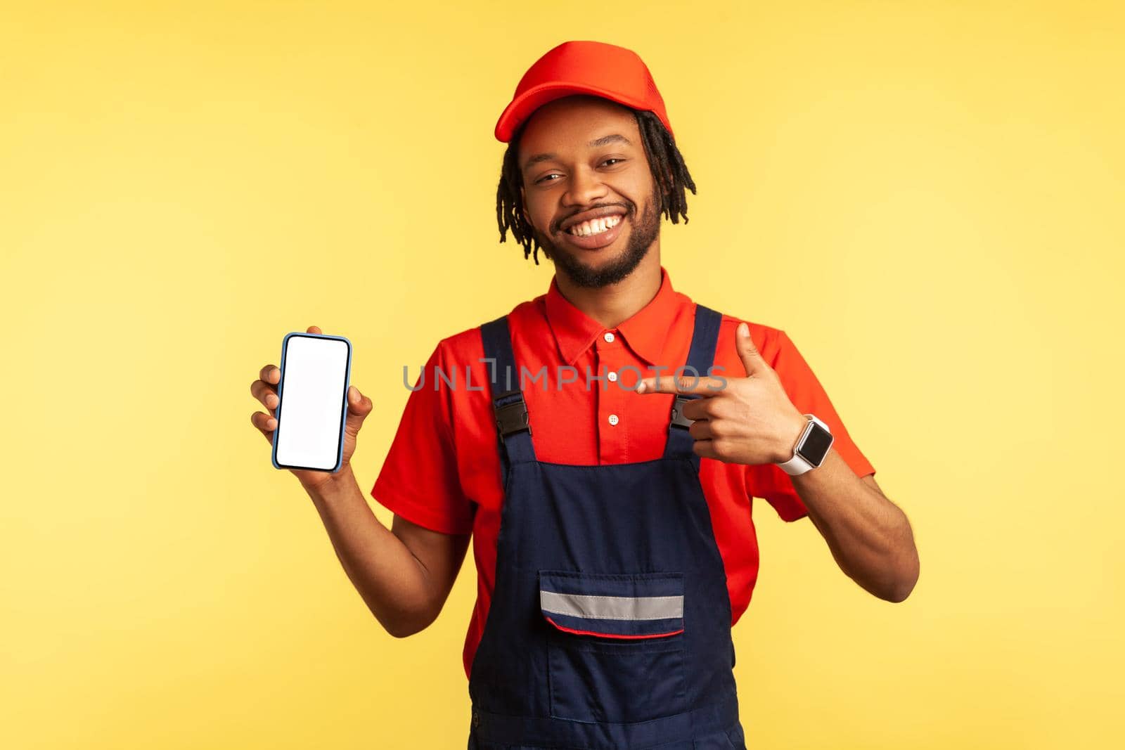 Portrait of happy handyman wearing uniform pointing at smart phone with blank screen, advertising new service industry application. Indoor studio shot isolated on yellow background.