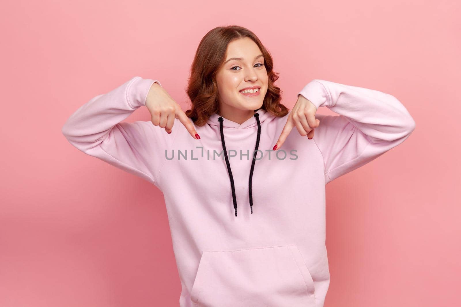 Portrait of excited teen curly haired female in hoodie pointing down place for commercial idea, looking at camera with toothy smile. Indoor studio shot isolated on pink background