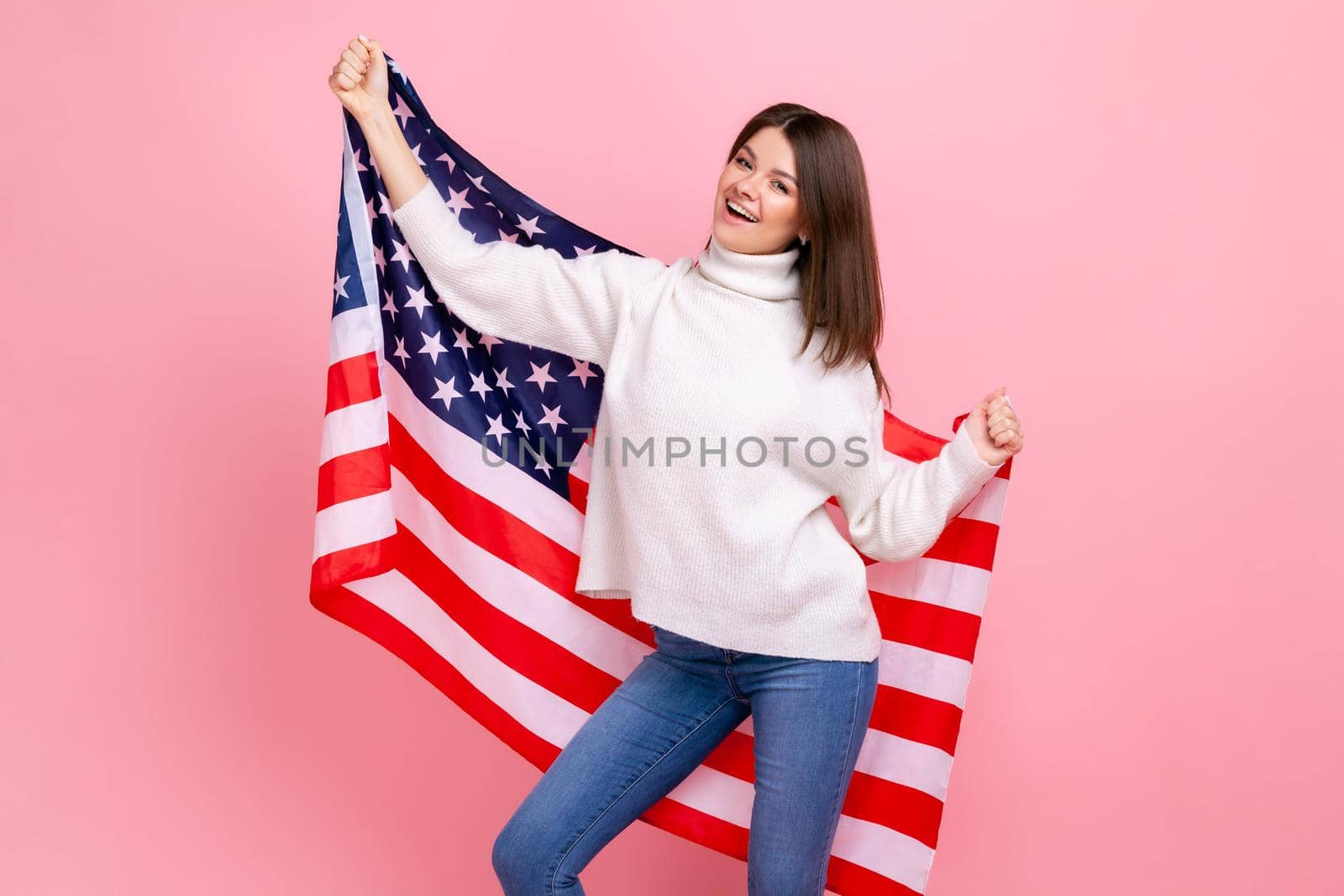 Portrait of pretty woman raised arms, holding american flag, celebrating national holiday, dancing, wearing white casual style sweater. Indoor studio shot isolated on pink background.