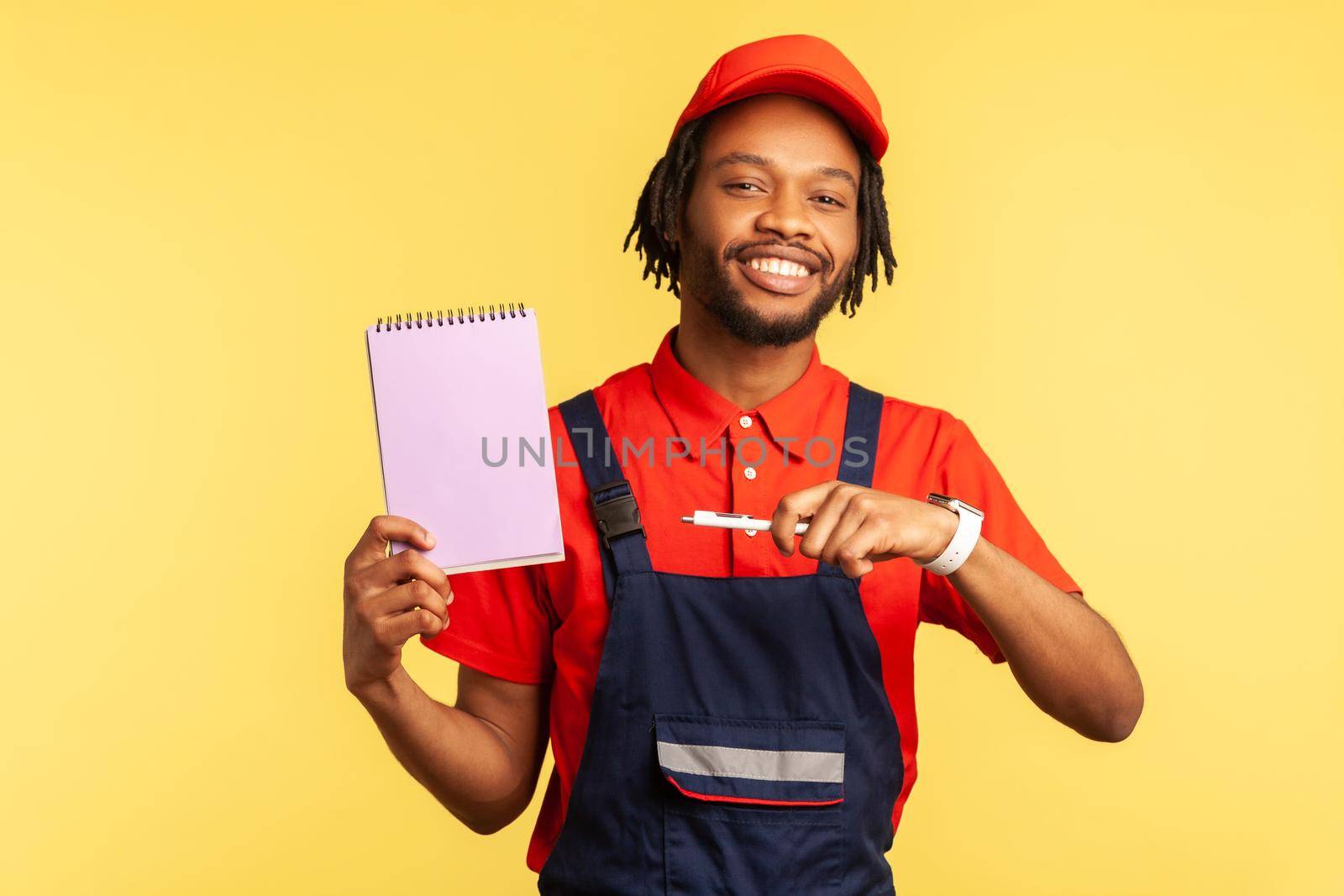 Portrait of workman wearing blue overalls and red cap, holding pointing at paper notebook with pleasant smile, presenting copy space. Indoor studio shot isolated on yellow background.