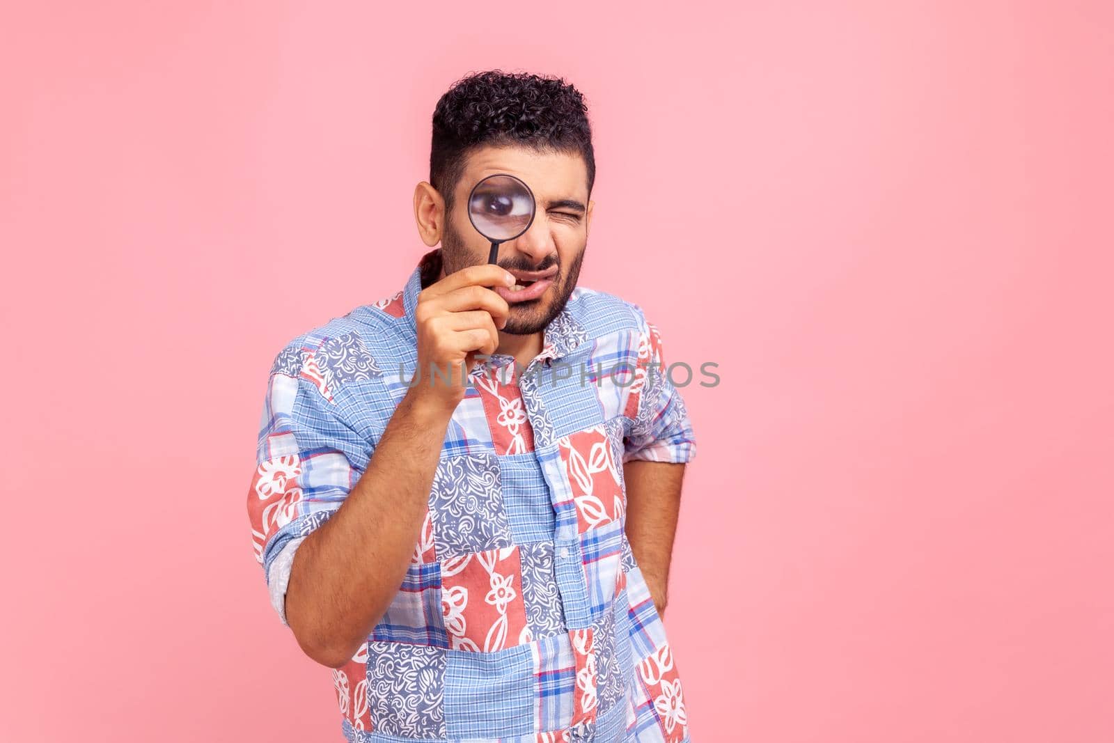 Bearded man in blue casual style shirt standing, holding magnifying glass and looking at camera with big zoom eye, having funny facial expression. Indoor studio shot isolated on pink background.