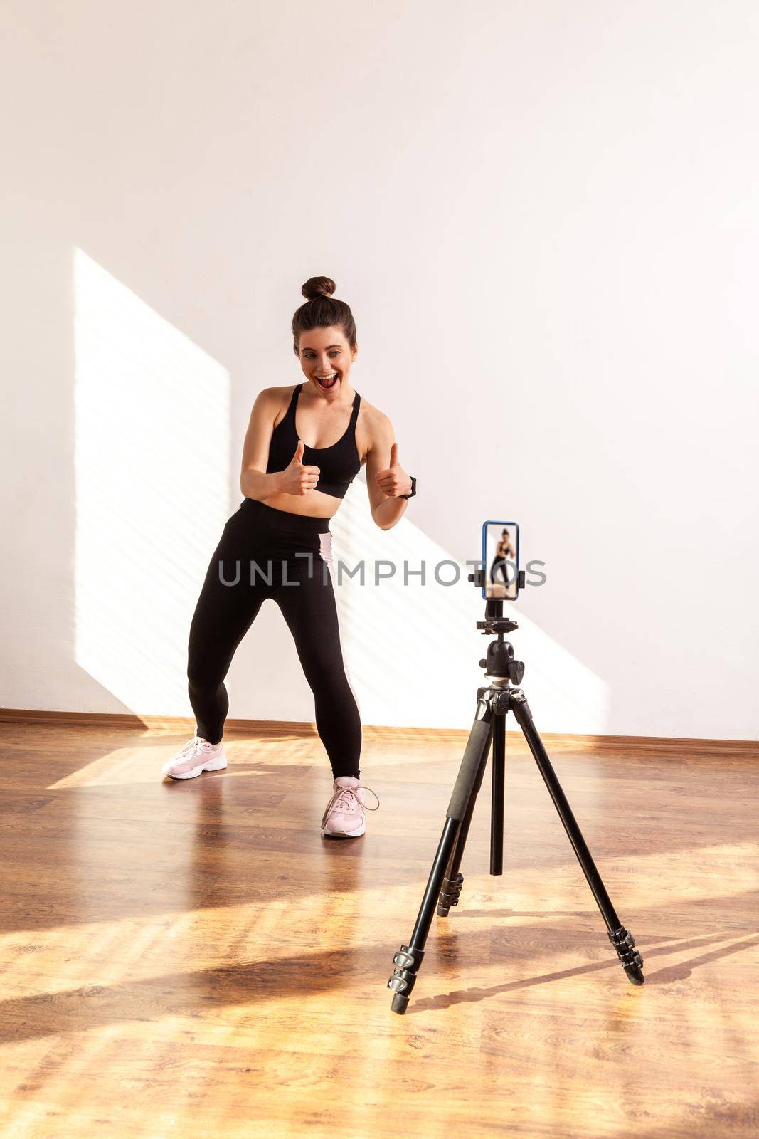 Fitness vlogger recording tutorials for sporty class, showing thumbs up to phone camera on tripod, wearing black sports top and tights. Full length studio shot illuminated by sunlight from window.