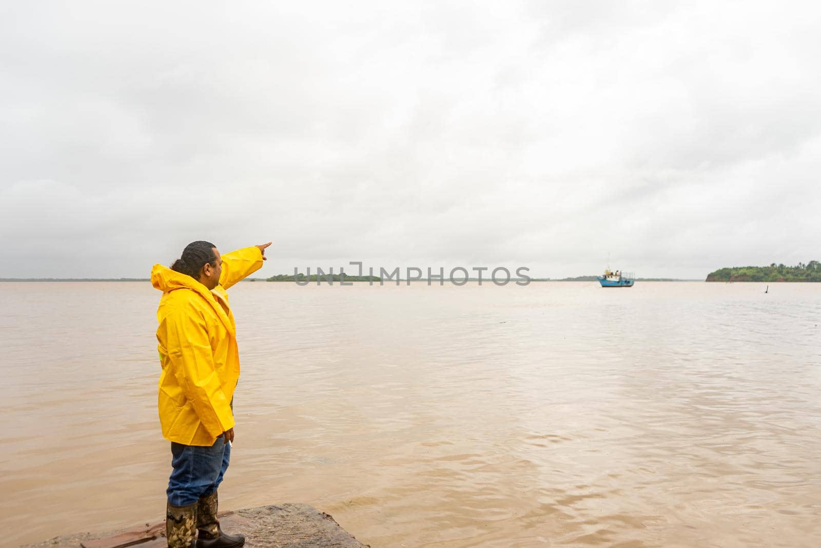 Latin mature man in yelllow raincoat pointing with his hand to a boat in the sea by cfalvarez