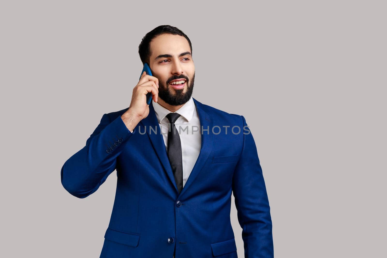 Smiling man standing, talking smartphone, making call and having pleasant conversation with friend or business partner, wearing official style suit. Indoor studio shot isolated on gray background.