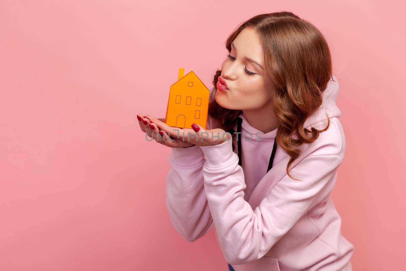 Portrait of joyful young brunette female in hoodie holding paper house in palms, looking with pout lips on model, dreaming about housing. Indoor studio shot isolated on pink background