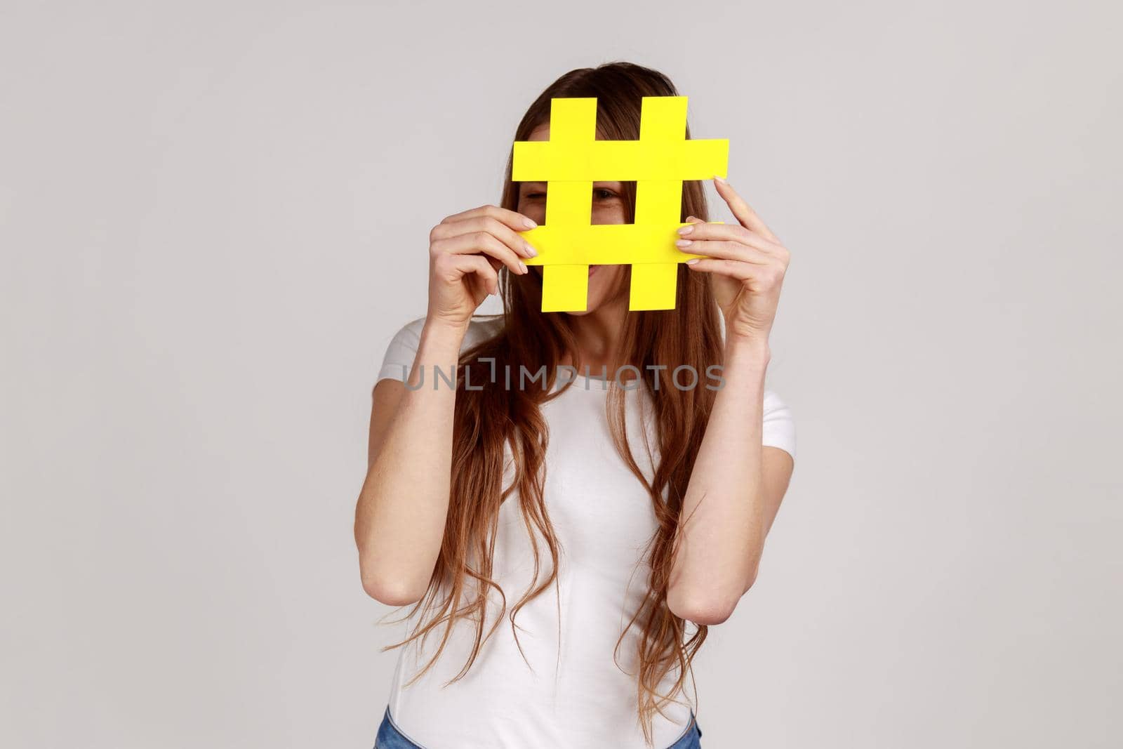Playful woman hiding behind yellow hashtag symbol, covering face with hash sign, concept of blogging and viral topics on internet, wearing white T-shirt. Indoor studio shot isolated on gray background