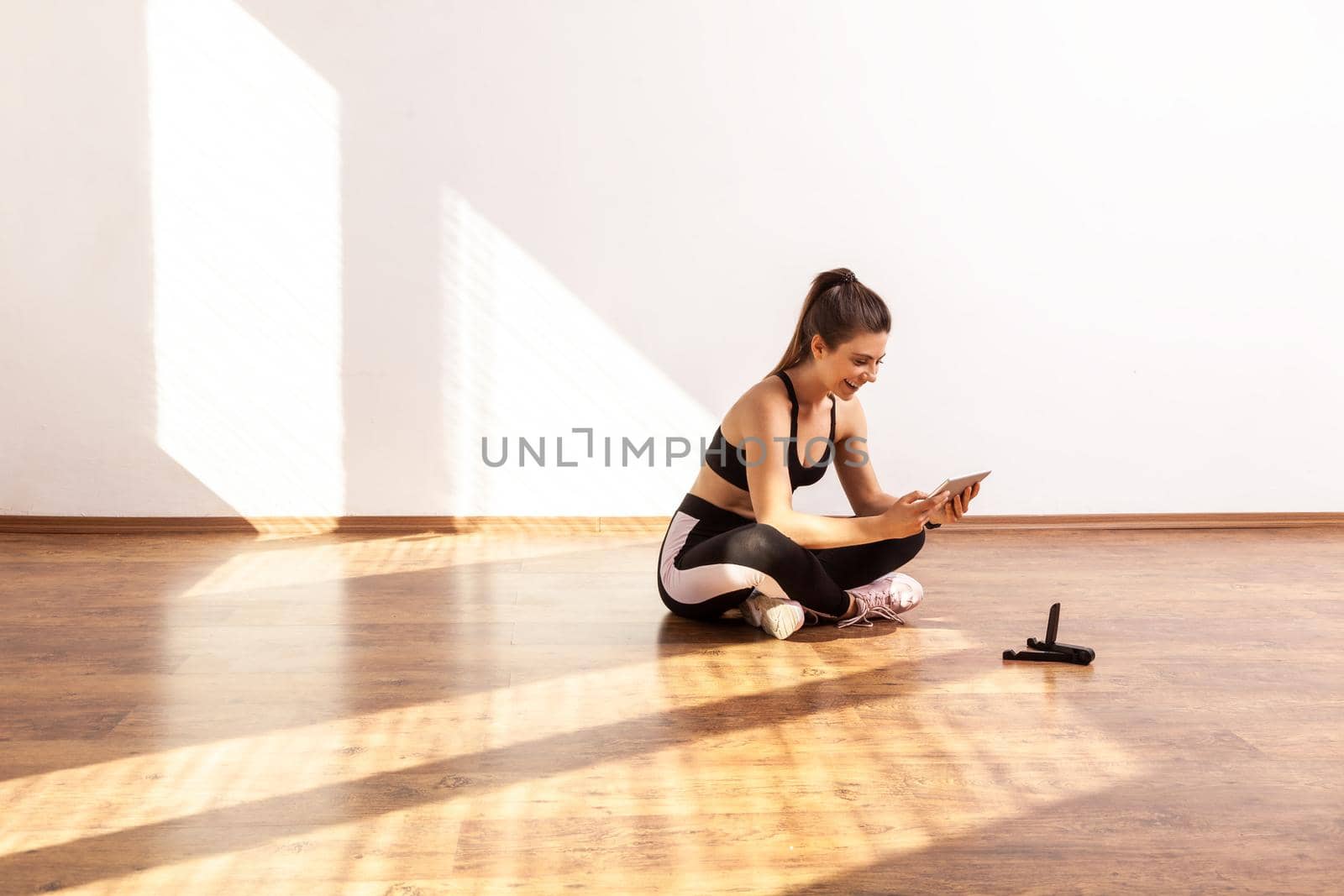 Girl sitting on floor with tablet, searching tutorial video for sport exercising, workout at home, wearing black sports top and tights. Full length studio shot illuminated by sunlight from window.