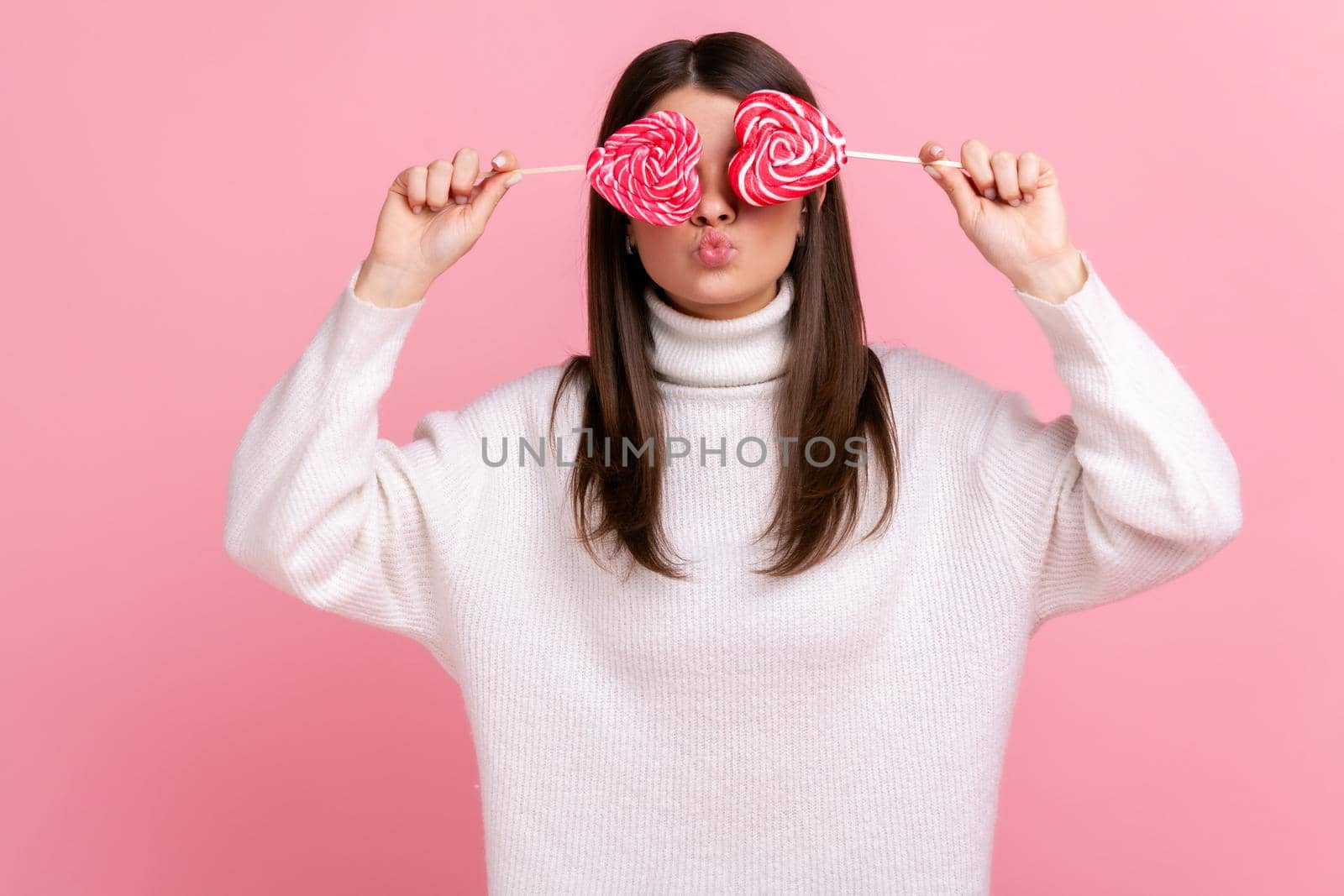 Portrait of playful childish romantic female covering eyes with sugary candies and sending air kisses, wearing white casual style sweater. Indoor studio shot isolated on pink background.