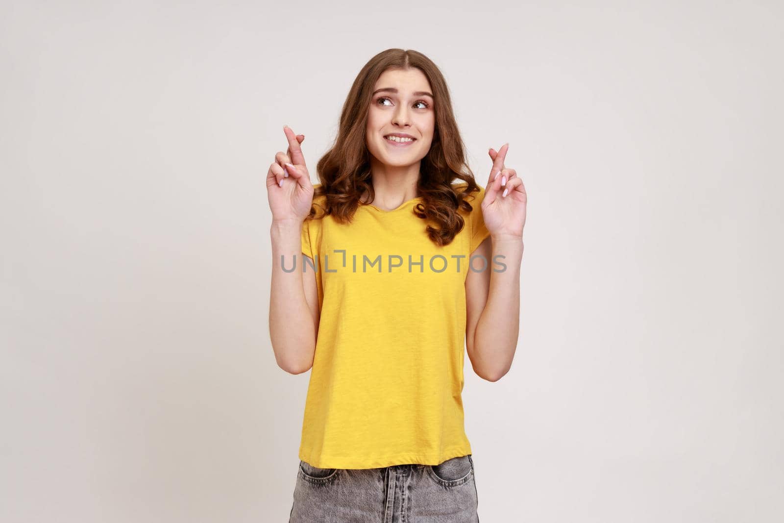 Hopeful nervous brown haired woman of young age in yellow casual T-shirt crossing fingers for luck, hope for better, ritual, looks up with hopeful look. Indoor studio shot isolated on gray background