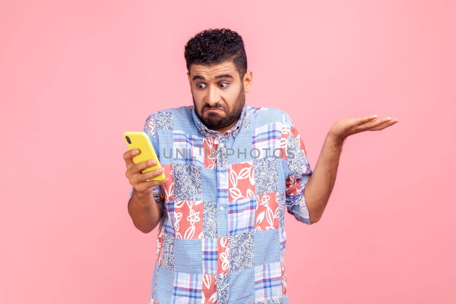 Portrait of attractive bearded man wearing blue casual shirt holding smart phone in hands, spreading hans aside, dont know how to use application. by Khosro1