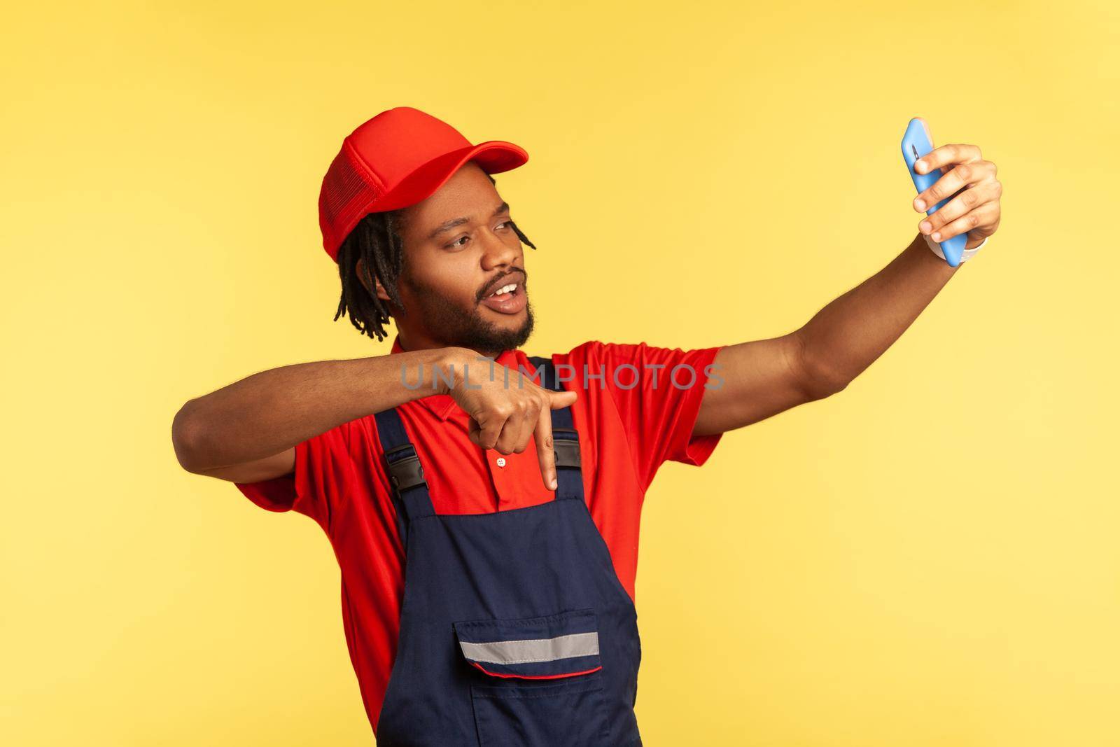 Handyman wearing blue uniform having video call with client, pointing down, asking to subscribe on social network of his service industry. Indoor studio shot isolated on yellow background.