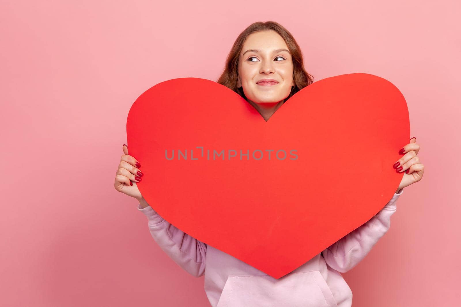 Portrait of happy teenage girl in hoodie holding huge paper heart and looking to side, falling in love, valentines day symbol. Indoor studio shot isolated on pink background