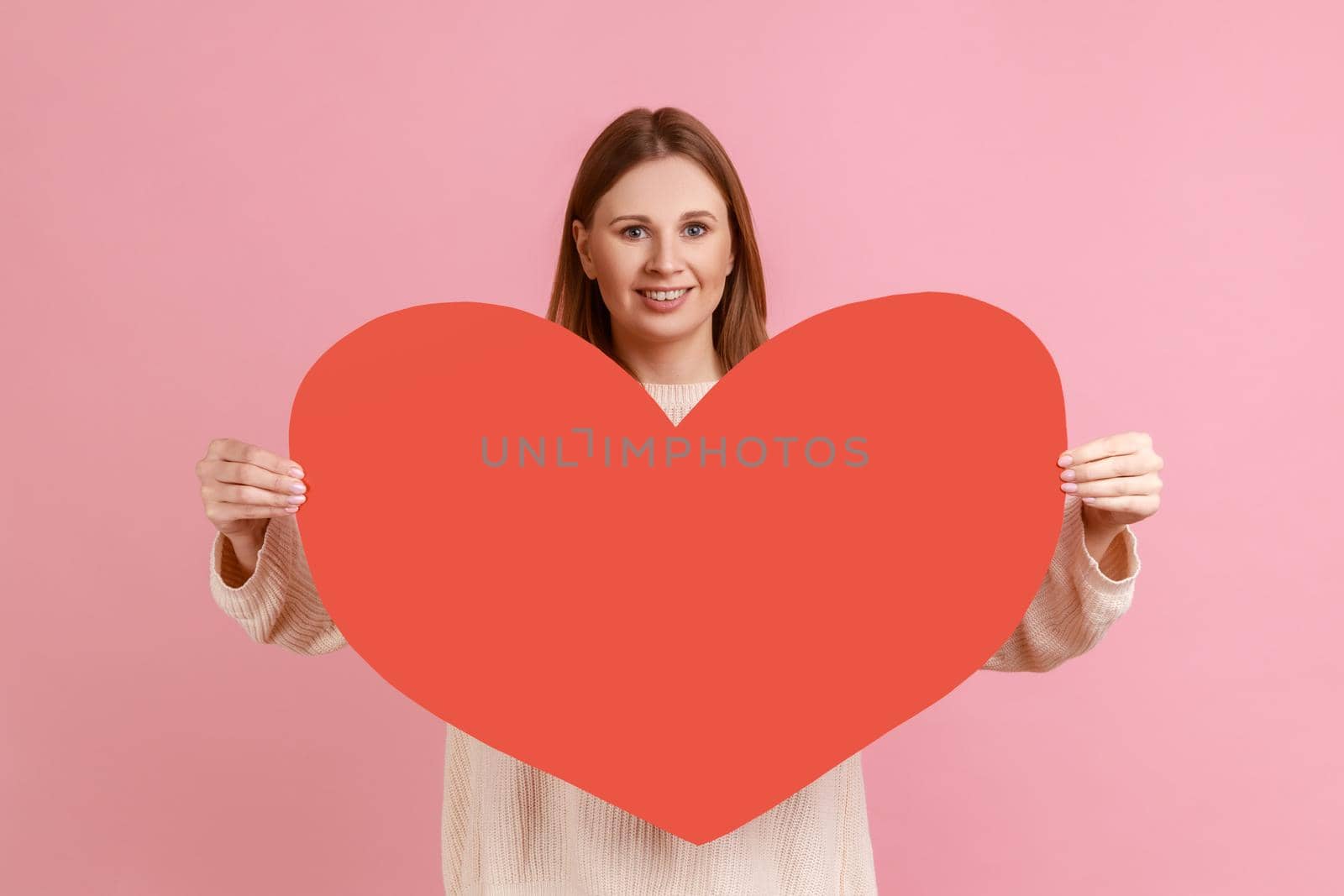 Portrait of happy romantic blond woman holding big red heart to camera and expressing love and positive emotions, wearing white sweater. Indoor studio shot isolated on pink background.