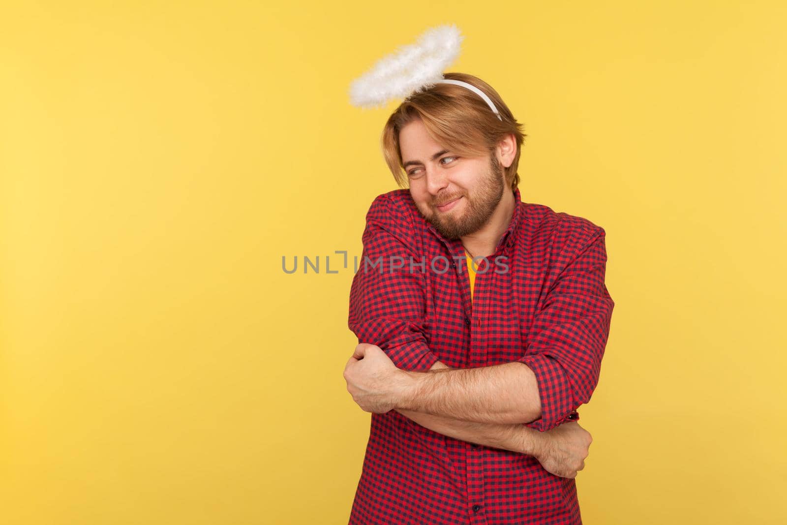 Portrait of hipster bearded guy in checkered shirt and with nimbus overhead hugging himself with love, worship to beauty, over-inflated ego. Indoor studio shot isolated on yellow background.