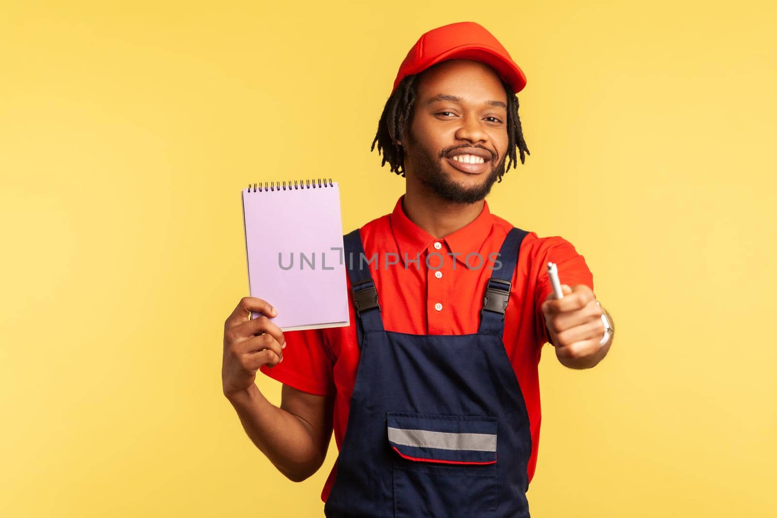 Portrait of optimistic mechanic or handyman wearing blue overalls and red T-shirt, showing paper notebook and holding out pen, copy space. Indoor studio shot isolated on yellow background.