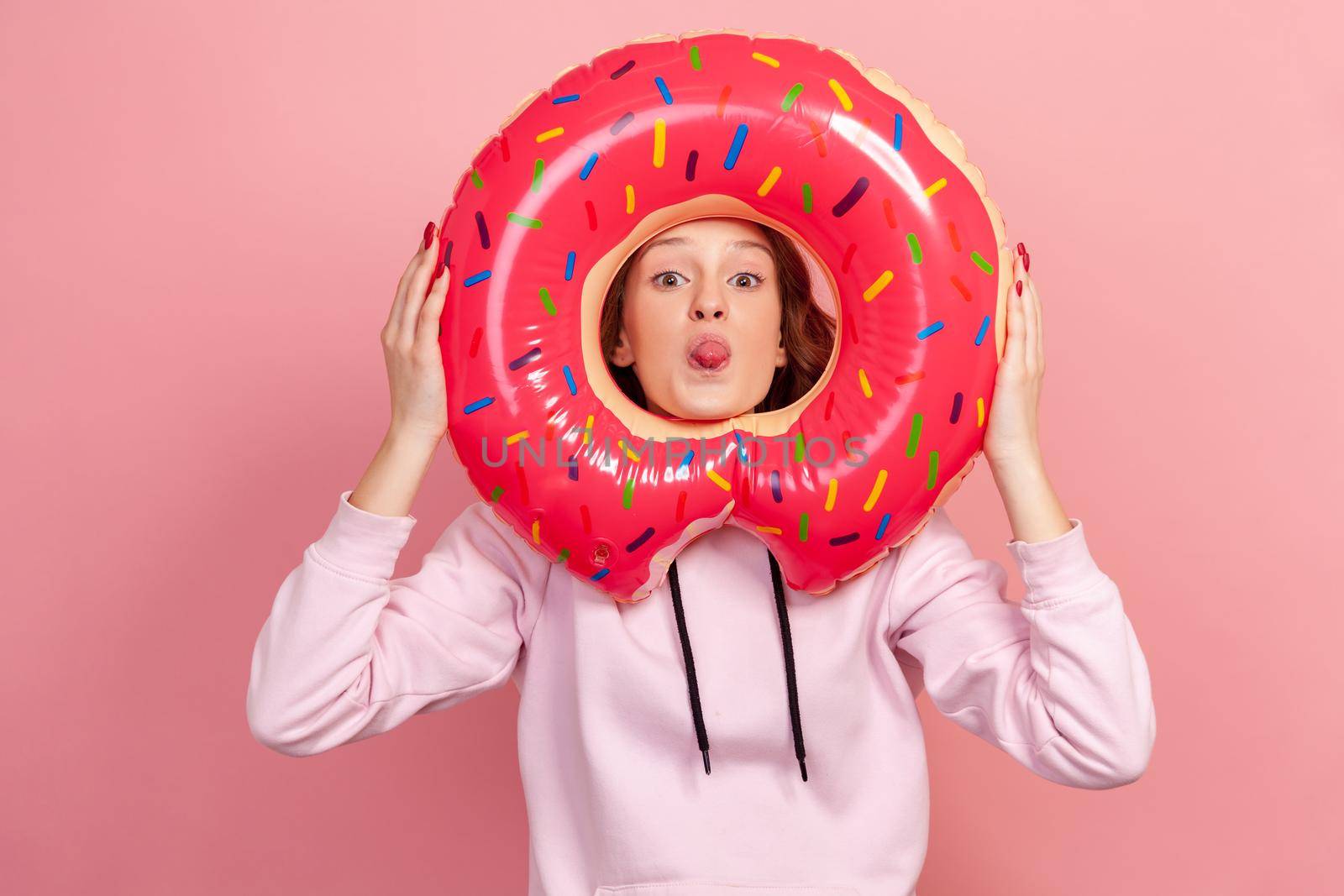 Portrait of funny curly haired teenage girl in hoodie showing tongue through pink donut rubber ring holding in hands, fooling around, rest. Indoor studio shot isolated on pink background