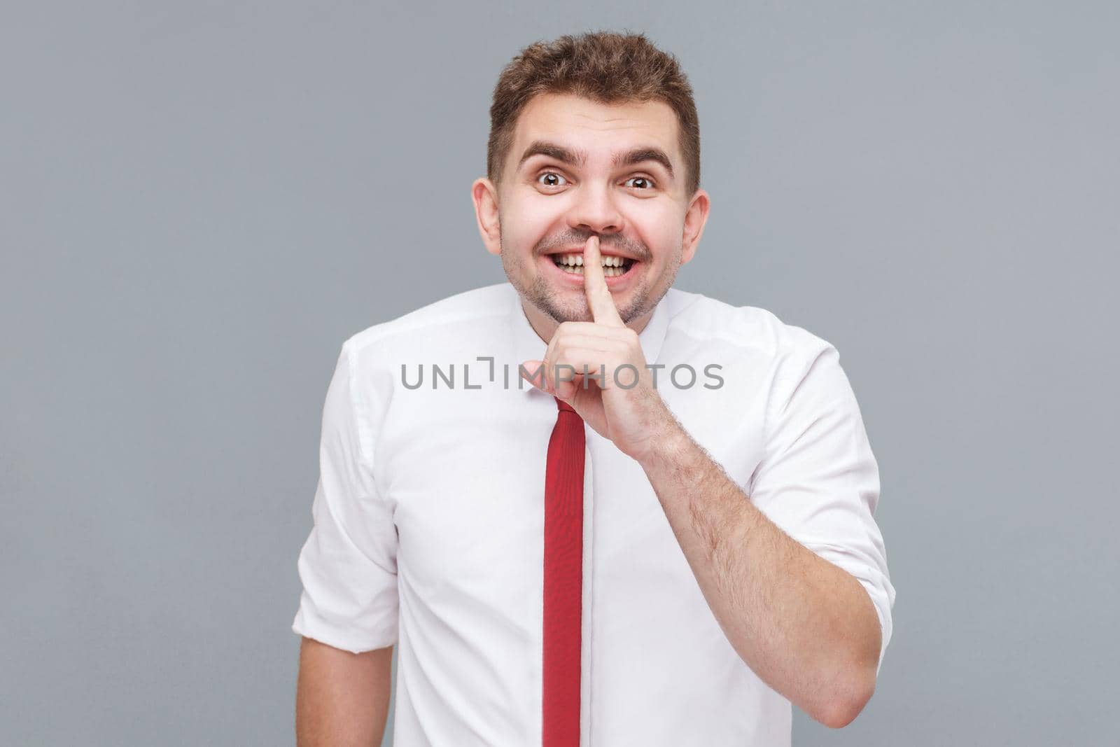 this is secret. Portrait of young handsome man in white shirt and tie standing with finger on lips, tell secret and smiling. indoor isolated on gray background.