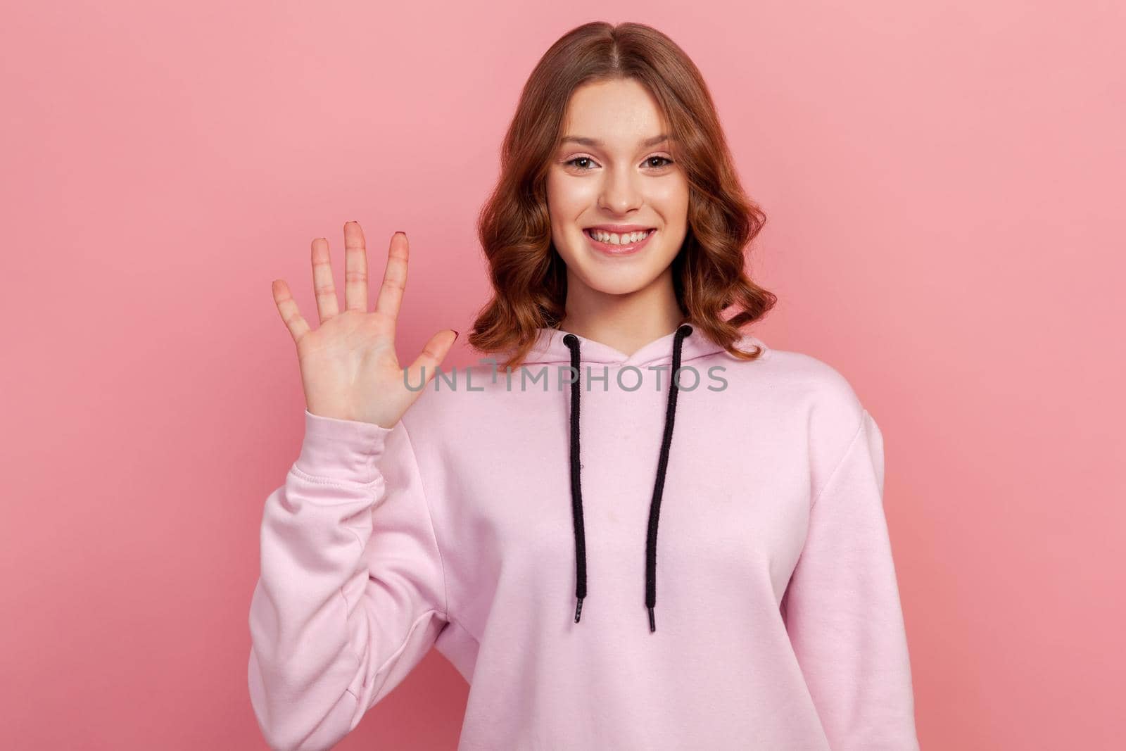Portrait of adorable friendly teen with brown hair in hoodie standing waving hand, looking at camera with engaging toothy smile, hello by Khosro1