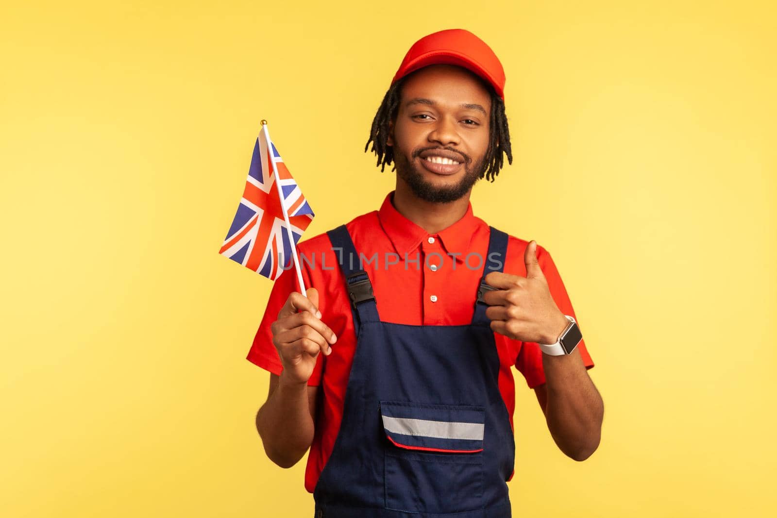Portrait of positive workman in blue overalls holding British flag and showing thumbs up, looking at camera with satisfied expression. Indoor studio shot isolated on yellow background.