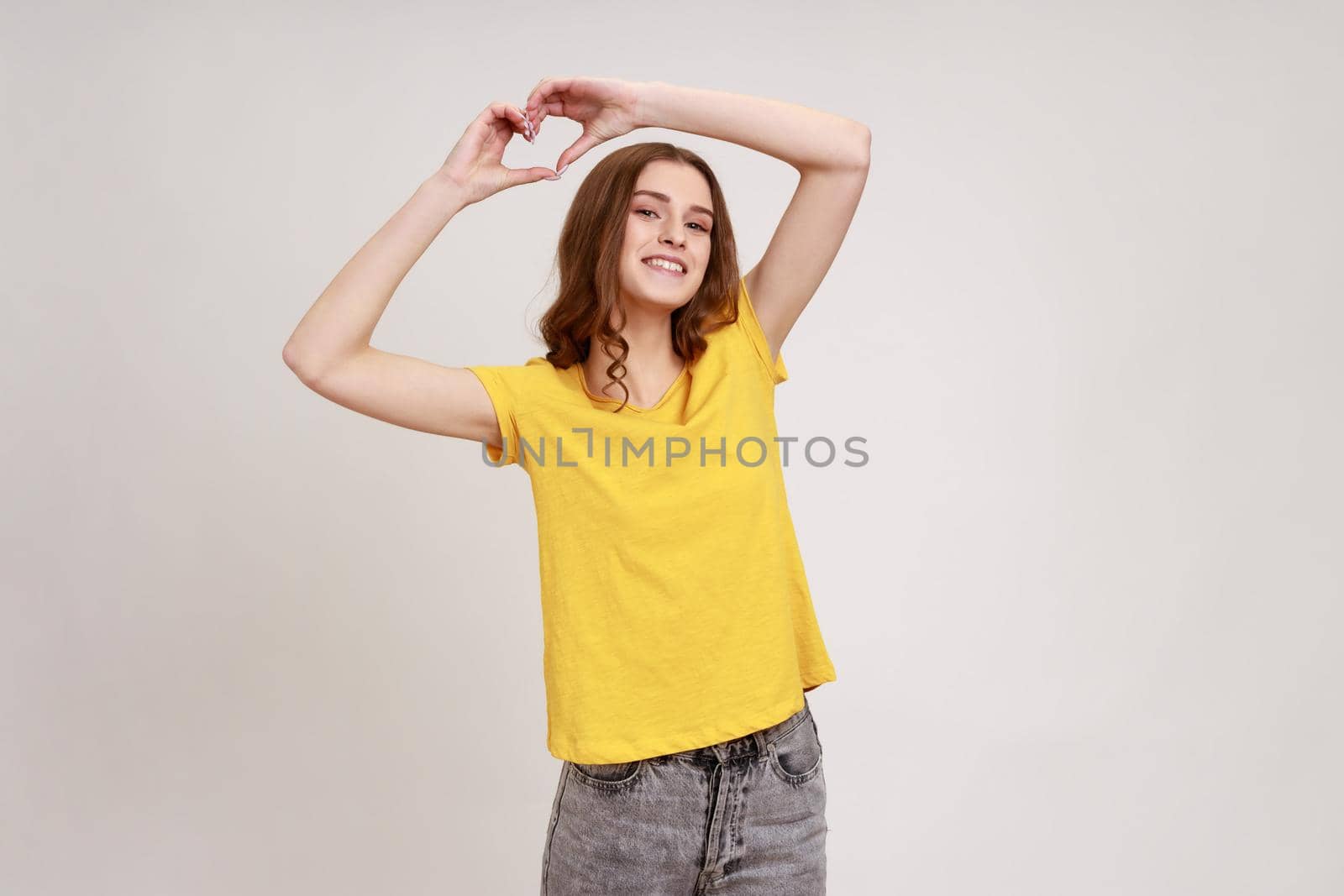 Happy beautiful teenager girl with brown wavy hair wearing T-shirt raising hands and showing heart shape gesture, flirting, demonstrating affection. by Khosro1