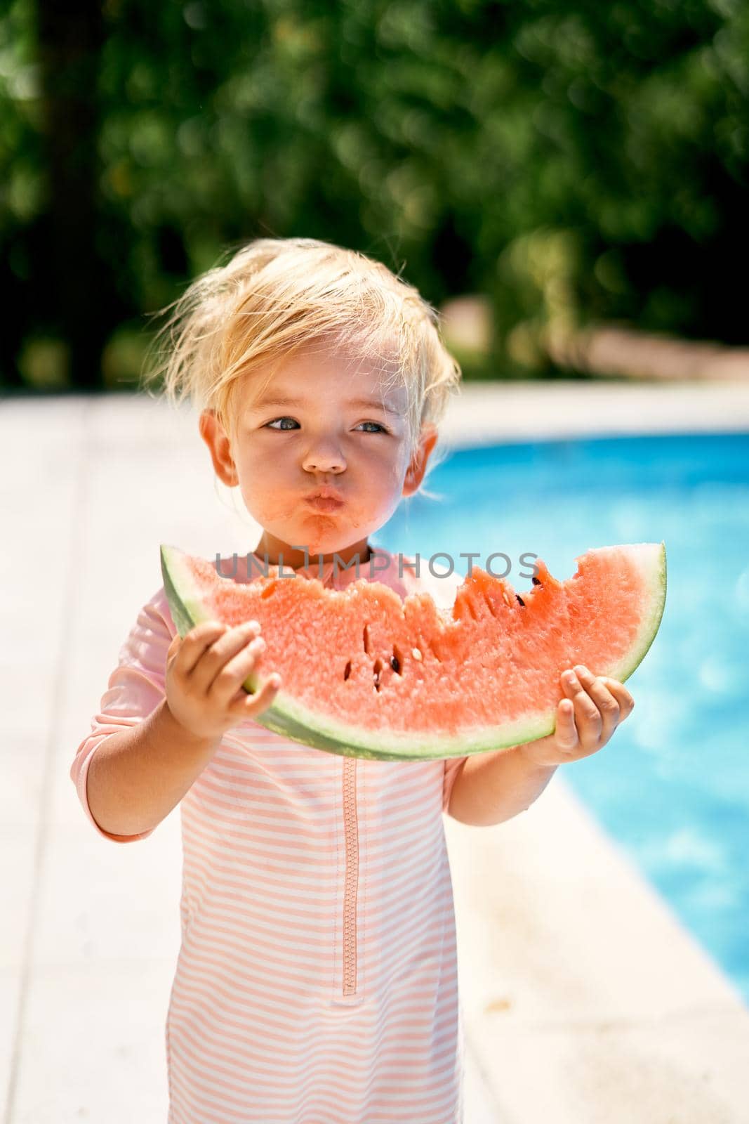 Little girl eats a watermelon near the pool with turquoise water by Nadtochiy