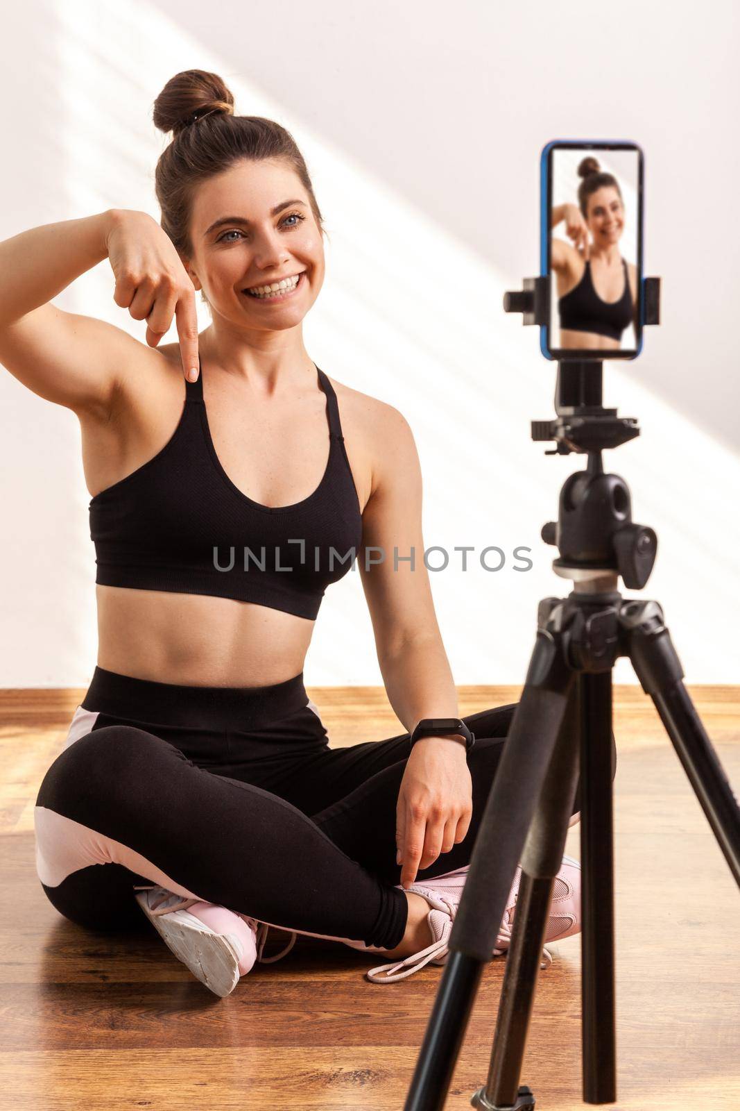 Online fitness trainer broadcasting workout online, points finger down, asking subscribe to her vlog, wearing black sports top and tights. Full length studio shot illuminated by sunlight from window.
