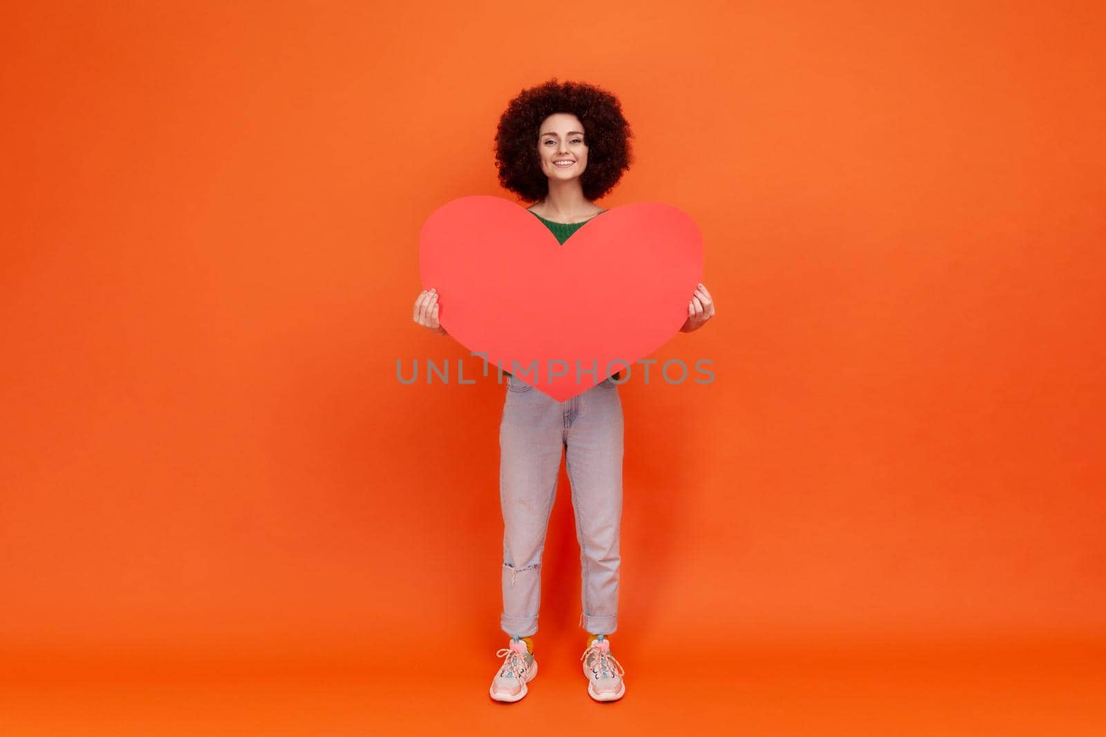 Full length portrait of falling in love woman with Afro hairstyle wearing green casual style sweater holding big red heart, looking with toothy smile. by Khosro1