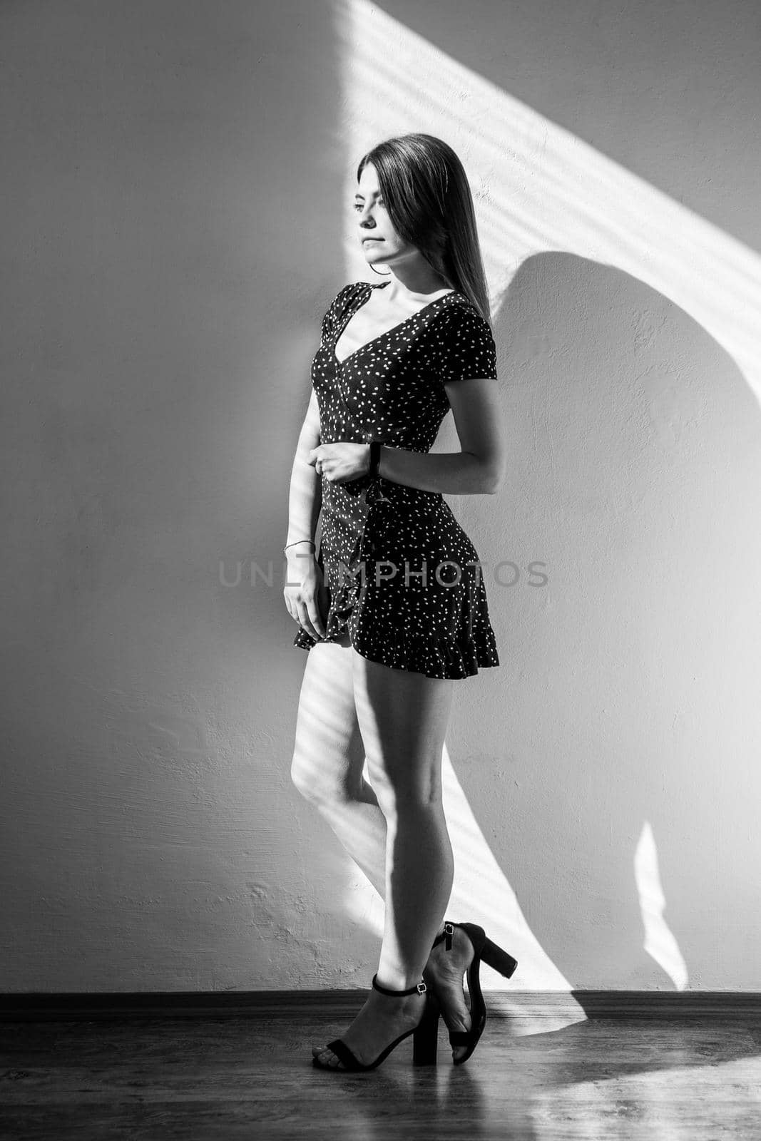 Attractive dark haired female standing looking away with romantic expression, wearing dress and sandals. Black and white photography, indoor studio shot illuminated by sunlight from window.