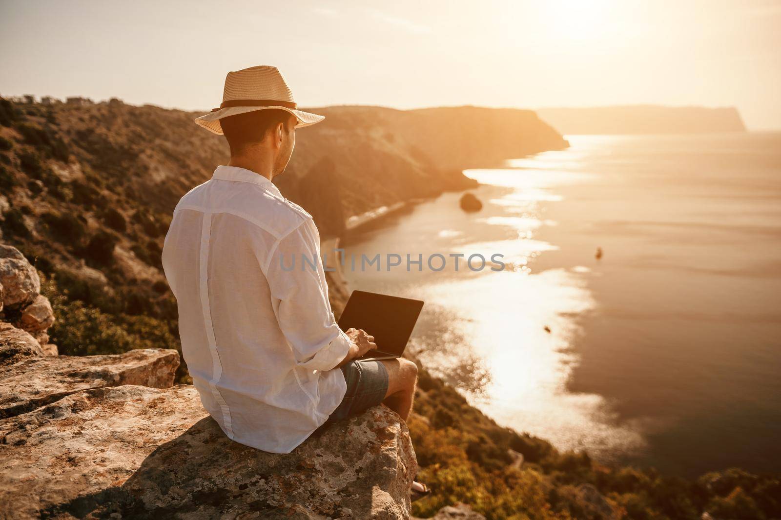 Digital nomad, freelancer. Remote work on vacation. Man in the hat, a businessman with a laptop sits on the rocks by the sea during sunset, makes a business transaction online from a distance. by panophotograph