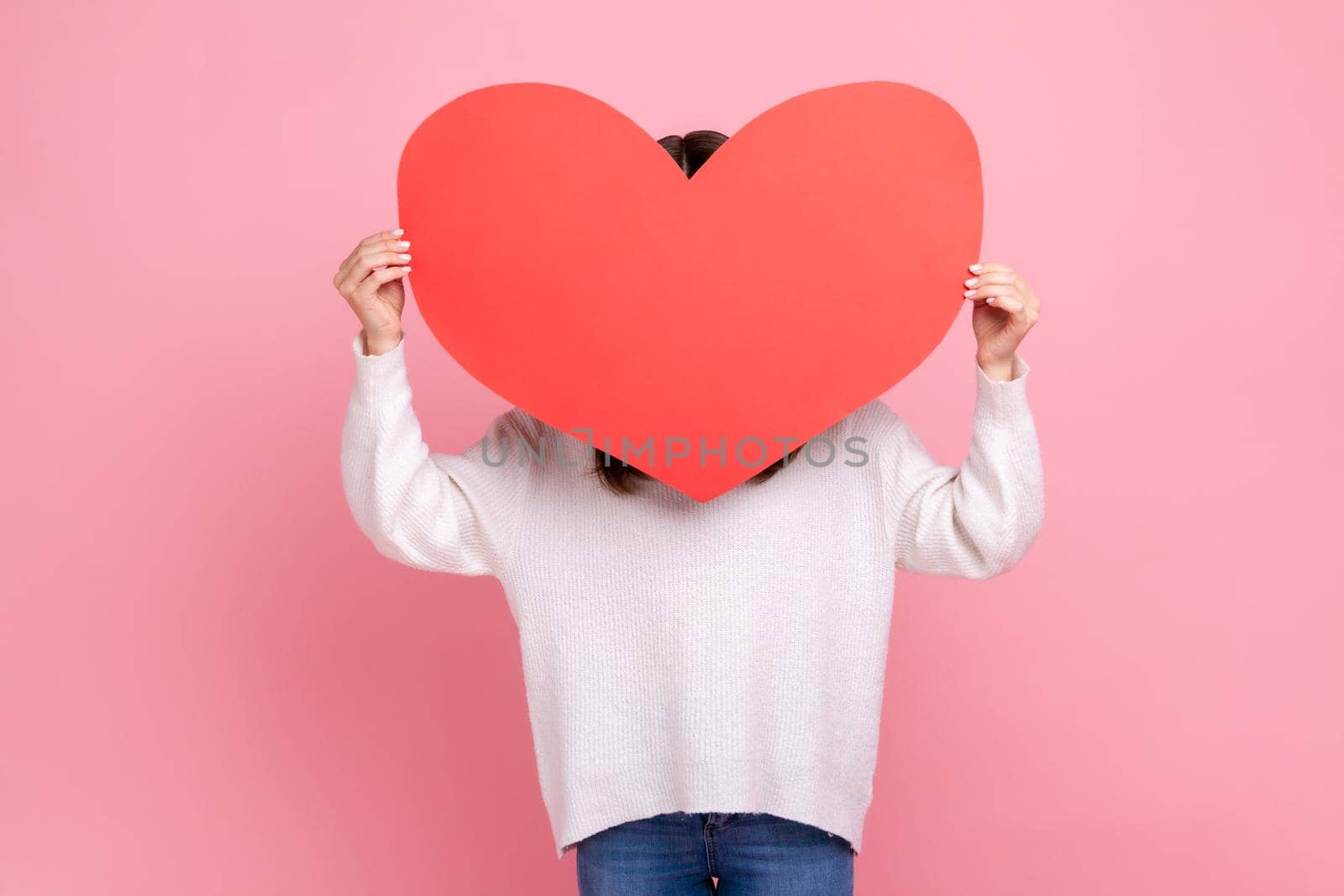 Unknown shy romantic woman covering her face with big red heart, hesitate to confess her feelings, wearing white casual style sweater. Indoor studio shot isolated on pink background.