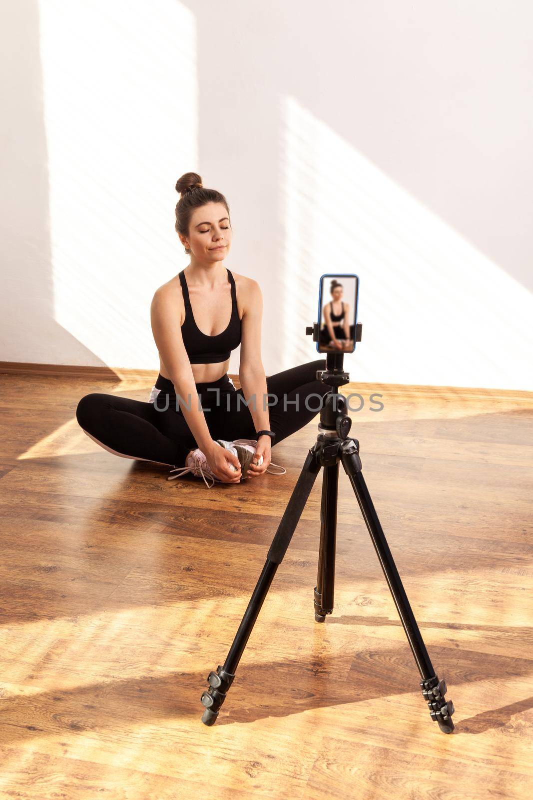 Girl recording online class of relaxing after training, sitting on lotus pose, keeps eyes closed, wearing black sports top and tights. Full length studio shot illuminated by sunlight from window.