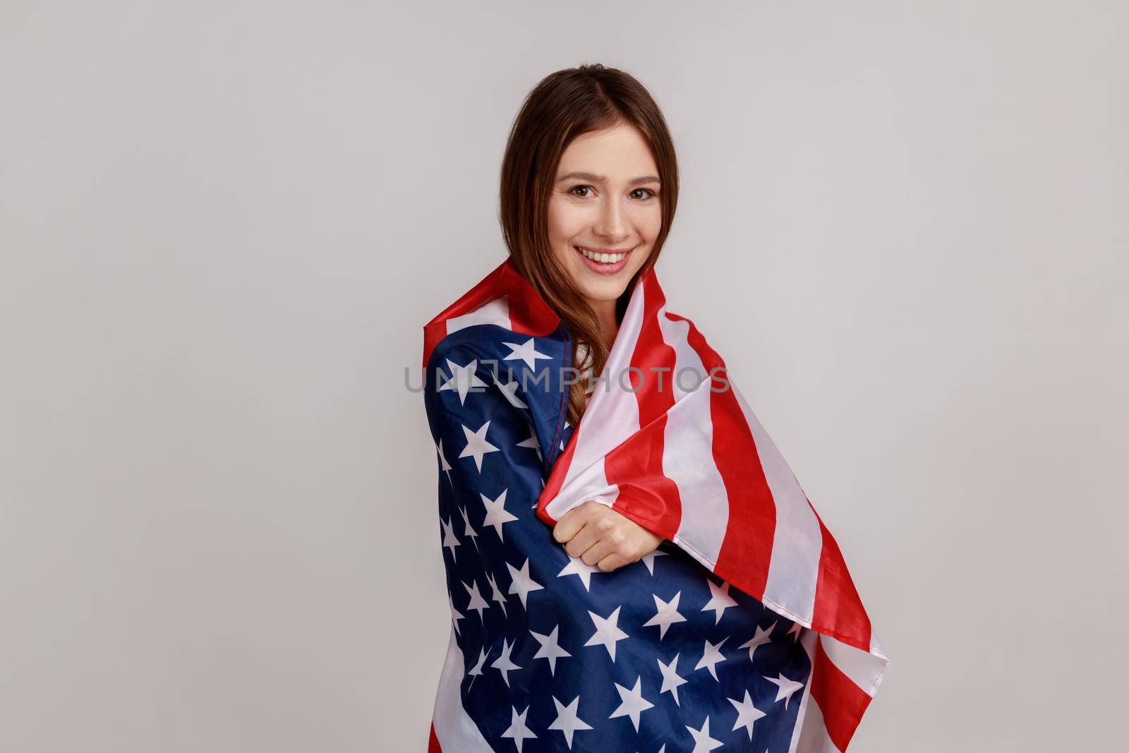 Portrait of friendly young woman standing wrapped in American flag looking at camera and smiling, wearing white casual style T-shirt. Indoor studio shot isolated on gray background.