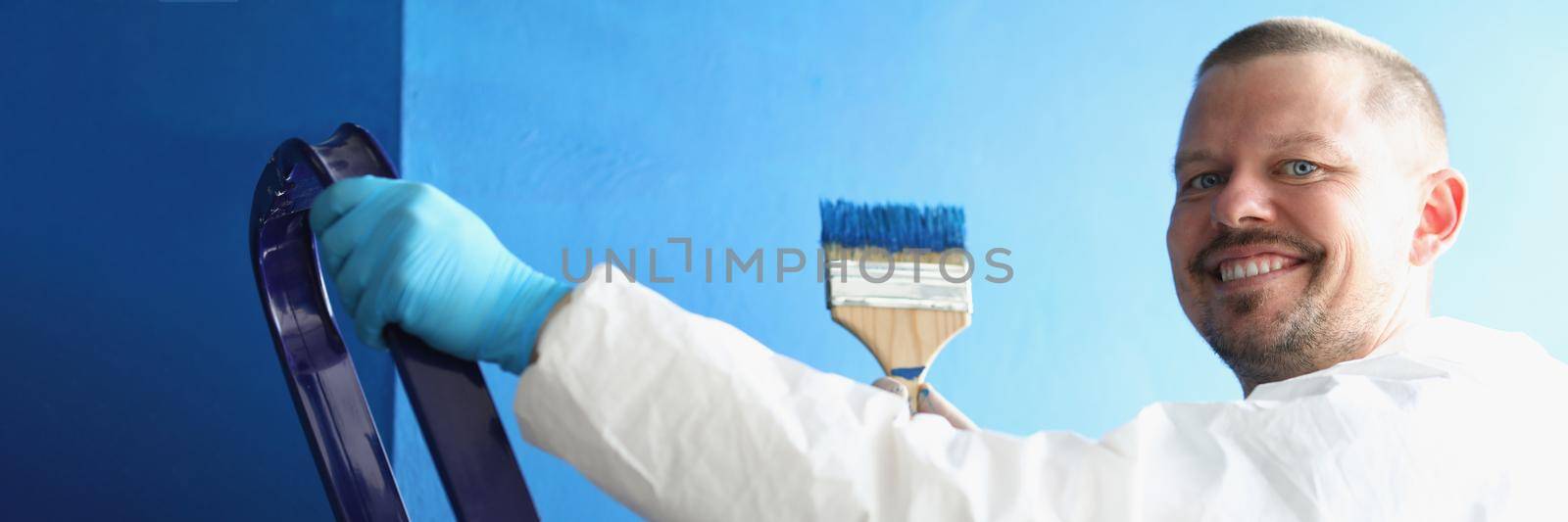 Portrait of painter holding brush covered in blue colour for painting walls, worker in uniform, happy and productive. Renovation, interior design concept