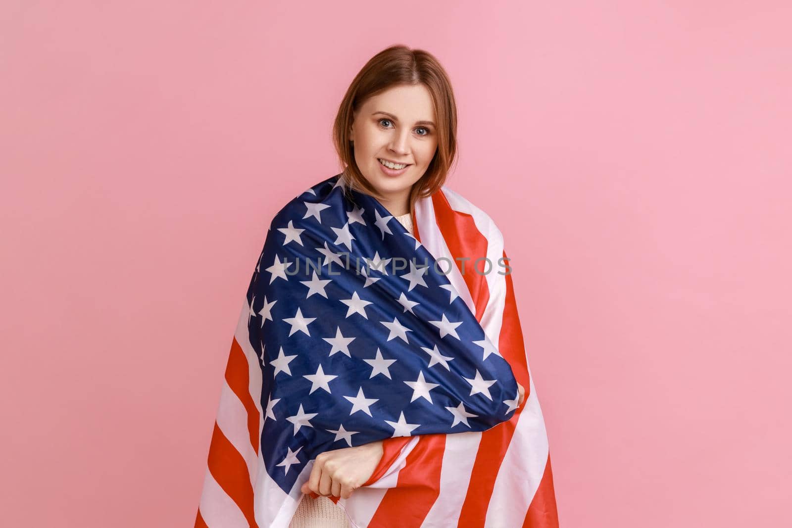 Portrait of kind friendly blond woman standing wrapped in american flag, looking at camera with pleasant smile, wearing white sweater. Indoor studio shot isolated on pink background.