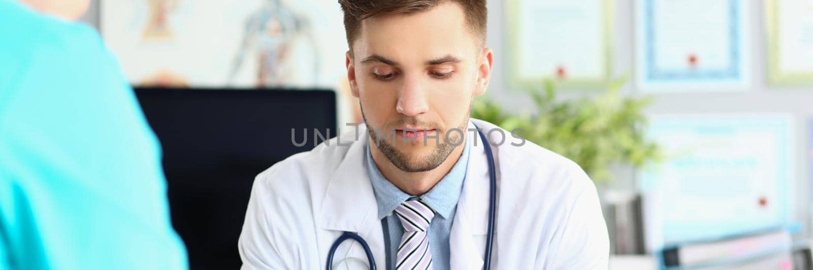 Portrait of young intern man in uniform and stethoscope, work meeting in doctor office. Listen to instruction of professional, make notes. Hospital concept
