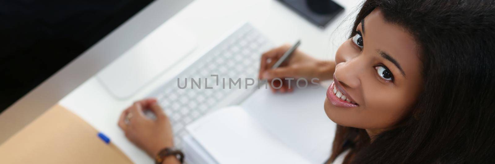 Smiling afro american young woman on workplace, open journal, luxury device on workplace by kuprevich