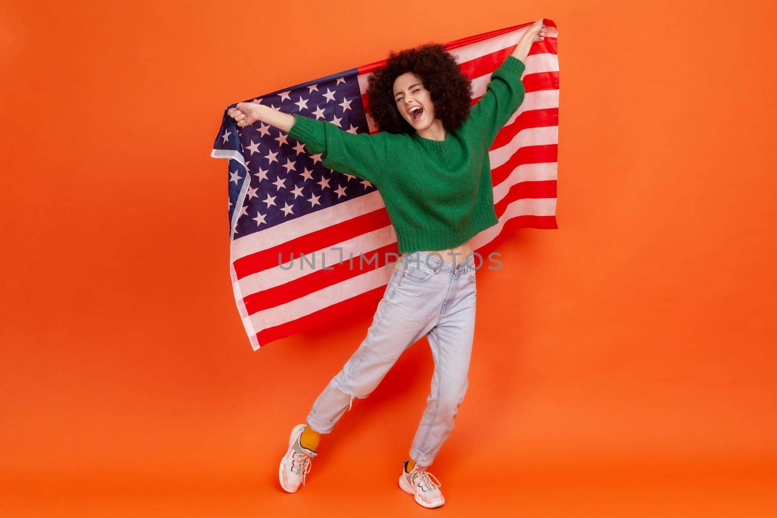 Full length portrait of happy woman with Afro hairstyle wearing green casual style pretending flying, having fun, holding american flag. Indoor studio shot isolated on orange background.