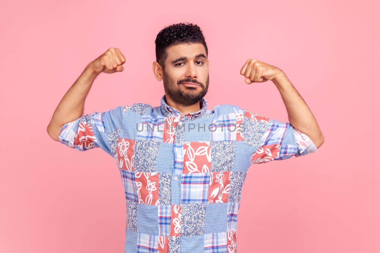 I'm strong. Portrait of attractive man in casual blue shirt raising hands to show biceps and looking confident at camera, feeling power and strength. Indoor studio shot isolated on pink background.