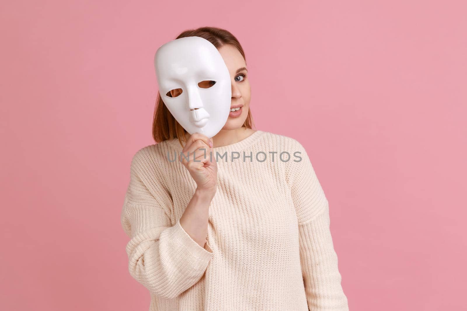 Portrait of smiling blond woman holding white mask, peeking, looking at camera, pretending to be another person, wearing white sweater. Indoor studio shot isolated on pink background.