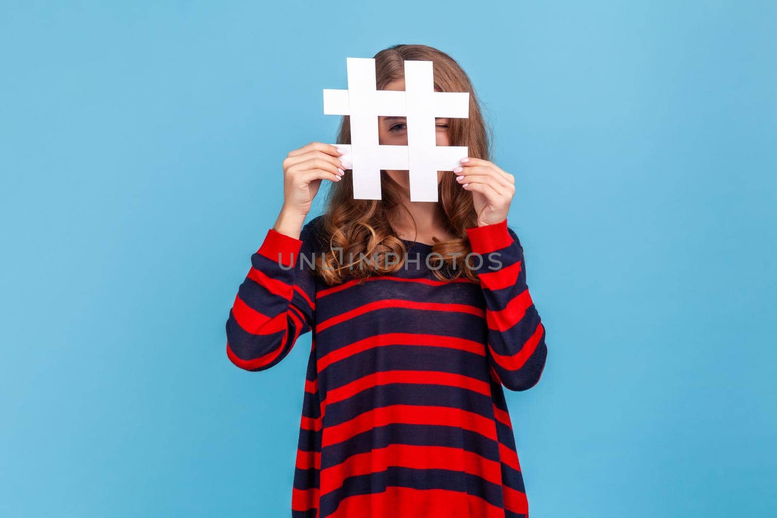 Curious woman wearing striped casual style sweater, looking at camera through large white hashtag symbol with prying eye, interesting web content. Indoor studio shot isolated on blue background.