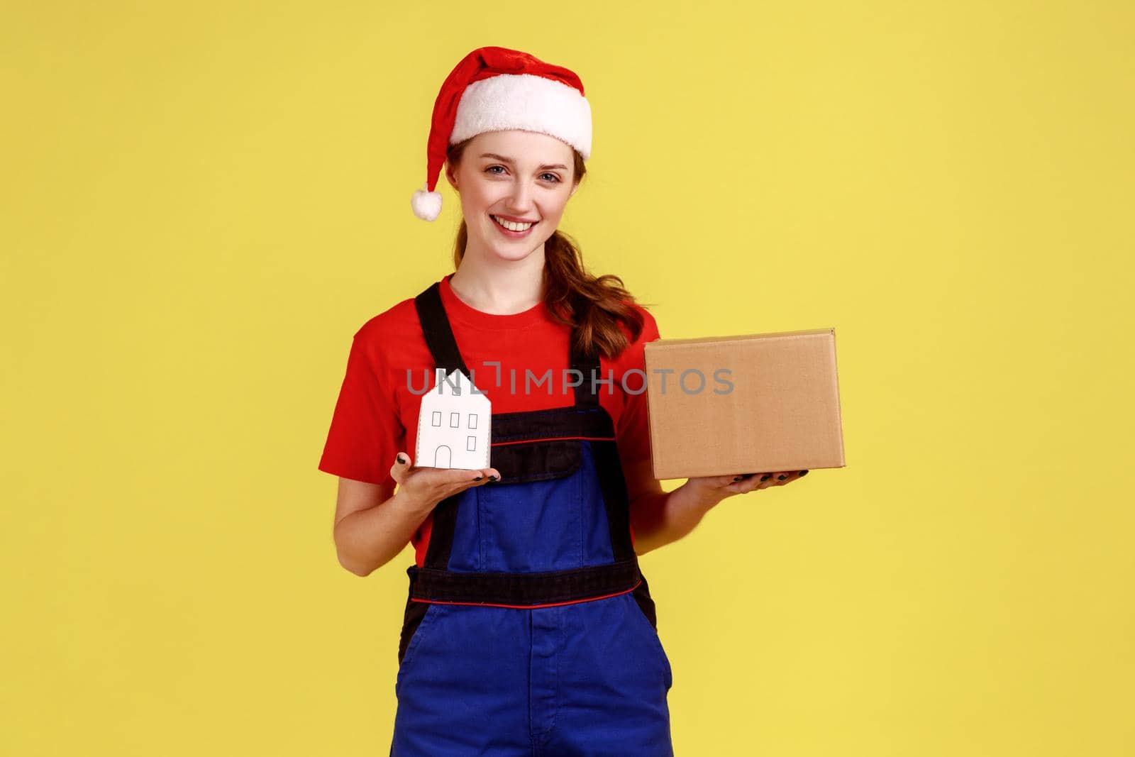 Smiling woman mover or worker holding cardboard parcel and paper house, relocation service, wearing blue overalls and santa claus hat. Indoor studio shot isolated on yellow background.