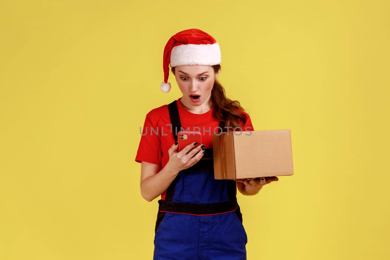 Surprised delivery woman with parcel in hands, using cell phone, having mistake with client address, wearing blue overalls and santa claus hat. Indoor studio shot isolated on yellow background.